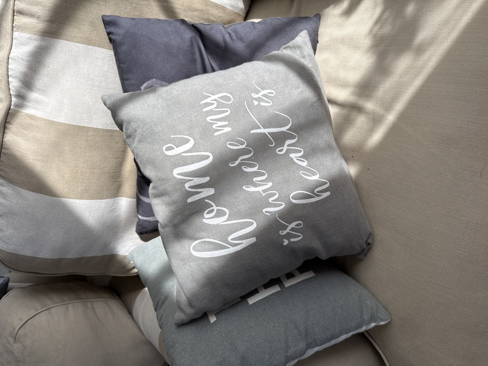 The image shows a close-up, high-angle shot of several decorative pillows placed on a beige sofa or chair. The pillows are arranged in a layered fashion, with the most prominent pillow in the foreground featuring white text that reads "home is where my heart is" in a cursive font. This pillow is a medium grey color. To its left is a lighter grey pillow with visible white text that appears to be "Th" followed by another letter. Behind these two pillows, a dark grey pillow is partially visible. In the background, a larger pillow with prominent beige and white vertical stripes can be seen. The lighting suggests the presence of sunlight filtering through blinds or trees, casting diagonal shadows across the scene. The image does not provide any specific information that would allow for its localization in Bad Sachsa, Germany.