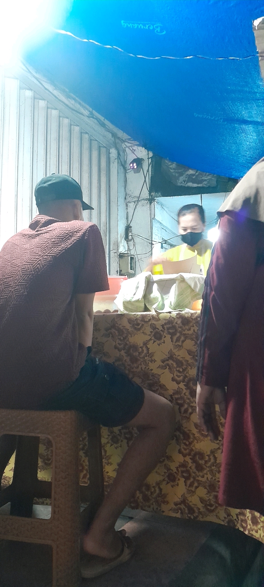 A low-angle, indoor shot captures a scene at what appears to be a food stall in Rogojampi, Indonesia. A person wearing a dark baseball cap and a maroon patterned short-sleeved shirt sits on a plastic stool, facing away from the camera and towards a counter. They are wearing dark shorts and sandals. To the right, partially obscuring the view of the person on the stool, is another individual wearing a dark red robe and a hijab. In the background, behind the counter, a person wearing a yellow shirt and a black face mask is actively serving customers or preparing items. The counter is covered with a yellow floral tablecloth. Several white packages, possibly food items, are visible on the counter. Above the scene, a bright blue tarp is stretched out, creating a canopy. The environment appears to be utilitarian, with corrugated metal siding visible on the left and electrical wiring and a light fixture in the upper right. The lighting is dim, with a bright light source likely originating from above and to the left, casting strong highlights and shadows.