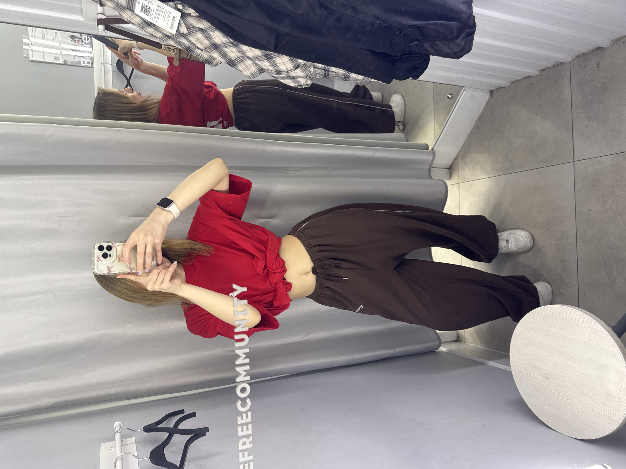 A young woman in a red tied shirt and brown sweatpants is taking a selfie in a fitting room. The room has grey curtains and a mirror showing a reflection of the woman. Clothes hang on a rack overhead. A white round stool is beside the woman. The walls are grey and the floor is grey tile. "FREECOMMUNITY" is printed on the left side of the image. There is no visible evidence to indicate that this photo was taken in Rostov-on-Don, Russia.