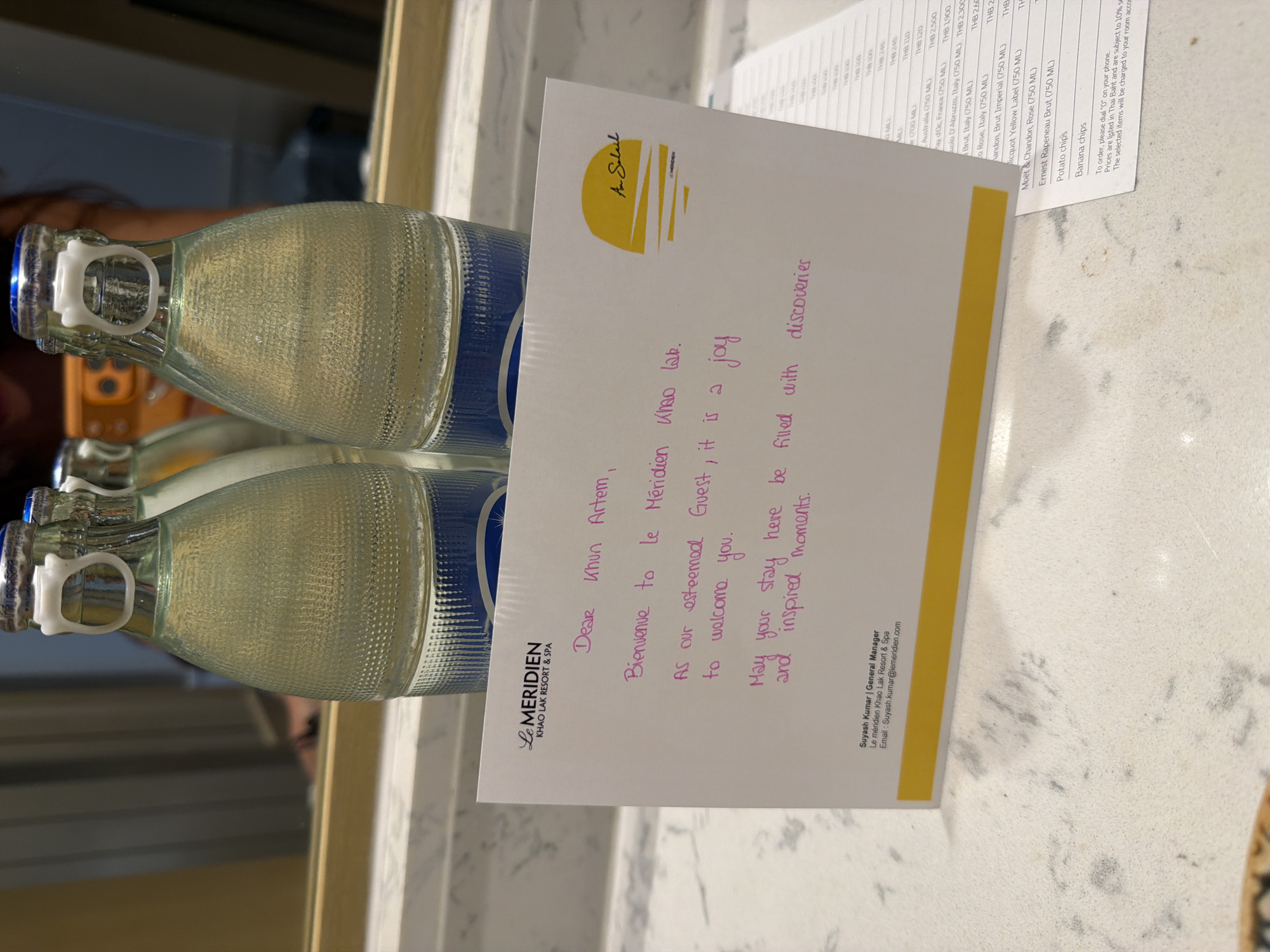 A handwritten note on a white card is placed on a white countertop, with two bottles of water in the background. The card, with the Le Méridien Khao Lak Resort & Spa logo, features a handwritten welcome message in pink ink. The message reads: "Dear Khun Artem, Bienvenue to le Méridien Khao Lak. As our esteemed Guest, it is a joy to welcome you. May your stay here be filled with discoveries and inspired moments." Below the message, "Suyash Kumar | General Manager" and "Le meridien Khao Lak Resort & Spa" are printed, along with an email address. To the right of the card, a portion of a menu or bill is visible, listing various items and prices in Thai Baht. The countertop is speckled white, with faint grey and black flecks. The background shows two clear glass bottles with blue labels, each containing water and sealed with a silver cap and white plastic closure. The bottles are positioned in front of a reflective surface, likely a mirror, which shows a blurred reflection of a person. The setting appears to be a hotel room, specifically in Bang Muang, Thailand, given the resort's location.