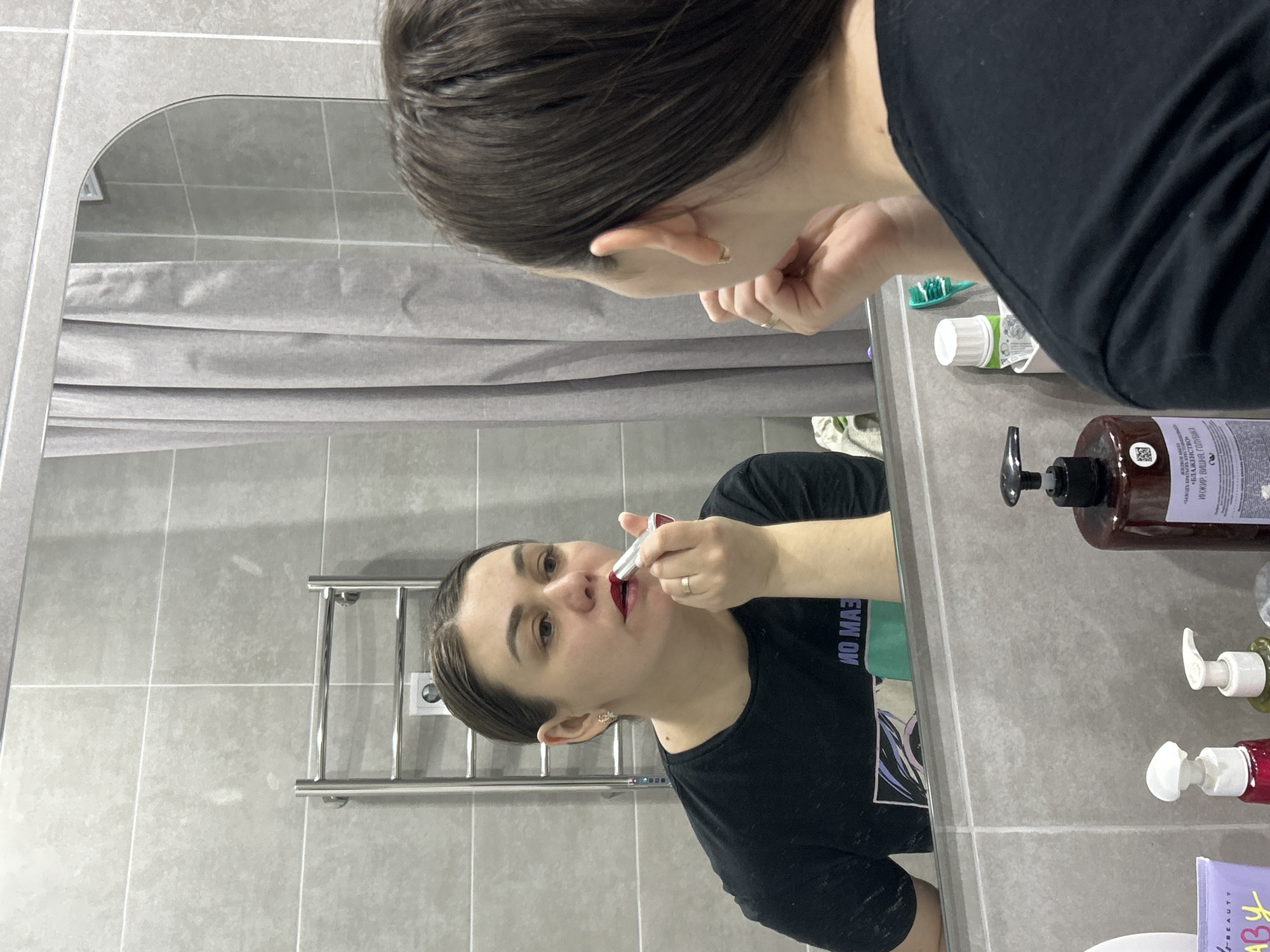 The image captures a woman in a modern bathroom in Chaikovsky, Russia, applying red lipstick while looking into a mirror. The photograph is oriented sideways, with the woman and her reflection appearing on their sides.
The woman has dark hair pulled back and is wearing a black t-shirt. Her reflection in the mirror shows her holding a silver lipstick tube to her mouth, concentrating on the application. Parts of her actual head and shoulder are visible outside the mirror's frame on the right and top.
The bathroom features light grey tiled walls and a grey fabric shower curtain in the background, both reflected in the mirror. A chrome heated towel rail is mounted on the wall to the left of the woman's reflection. On the grey countertop reflected in the mirror, various personal care items are visible, including a large brown pump bottle, a green toothbrush, a tube of toothpaste, and other smaller pump bottles and a purple pouch.
Visible text on the brown pump bottle includes "ИЖИР ВИШНЯ" (Fig Cherry) in Cyrillic, along with other unreadable Cyrillic text. The purple pouch has "BEAUTY" and "БАБУ" (Babu) partially visible. On the woman's black t-shirt, a mirror-reversed partial text, possibly "ИО МАЗ" (IO MAZ), can be discerned. The scene depicts a routine personal grooming moment in a clean, functional bathroom setting.