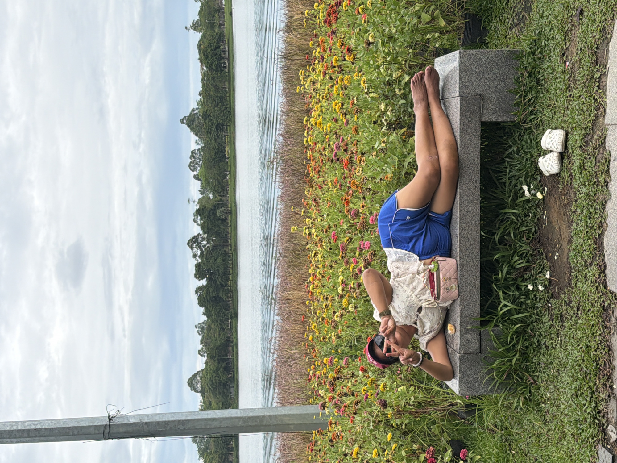 A person is lying on a granite bench in a field of colorful flowers, with a lake and trees in the background. The person is wearing a white top, blue shorts, sunglasses, and a red hat. They are making a peace sign with their fingers. There is a small pink bag on the bench next to them, and a pair of white sandals on the ground in front of the bench. The scene appears to be in Thuận Hóa Ward, Vietnam, based on the location information provided. The sky is cloudy, and the overall atmosphere is relaxed and peaceful.