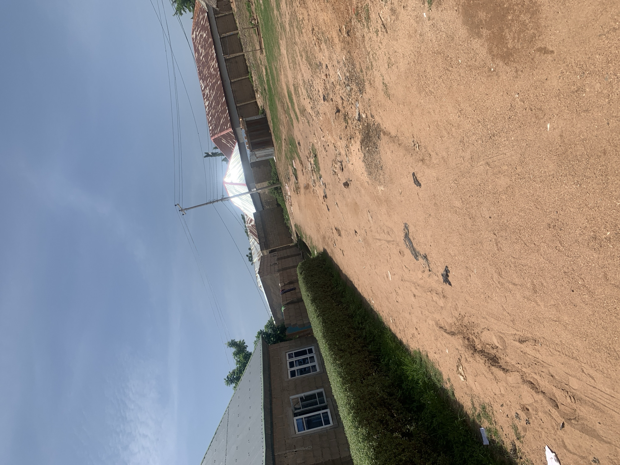 The image depicts a street scene in the city of Jauro Yinu, Nigeria. The foreground is dominated by a dirt road with visible tire tracks and scattered debris. To the left, a neatly trimmed green hedge runs along the side of a modest, light-colored brick building with white-framed windows. Beyond this building, other structures are visible, including houses with corrugated metal roofs, some showing signs of rust. A wooden fence separates some of the properties. Electrical poles and overhead wires are present, indicating the presence of utilities. The sky is a clear blue with a few wispy white clouds. The overall impression is that of a residential area in a warm, possibly dry climate, with simple architecture and unpaved roads. There are no people visible in the image.