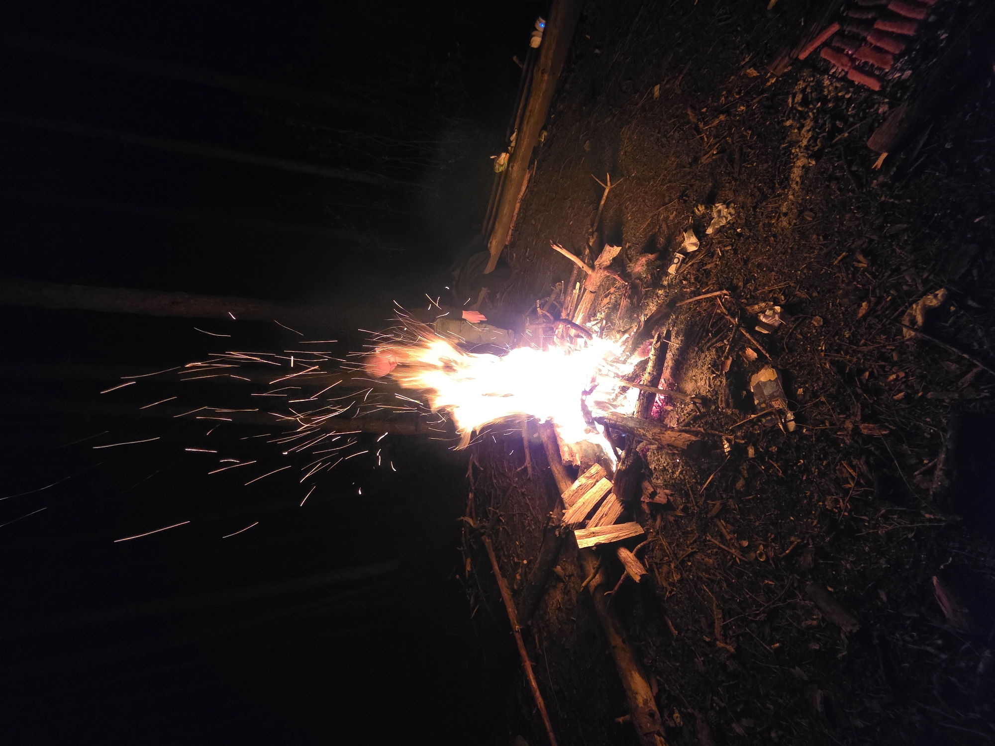The image captures a night scene in an unspecified location in the city of Unknown, Unknown, featuring a fire burning with intensity. The fire is constructed with wood and branches, emitting light and sparks that illuminate the surrounding darkness. The foreground consists of dirt, dry leaves, and small debris, along with some packages of fireworks. The environment suggests an outdoor setting, possibly a wooded area or backyard, with a wooden structure and additional objects visible in the background, though their details are not fully discernible due to the dim lighting. No people are visible in the frame.