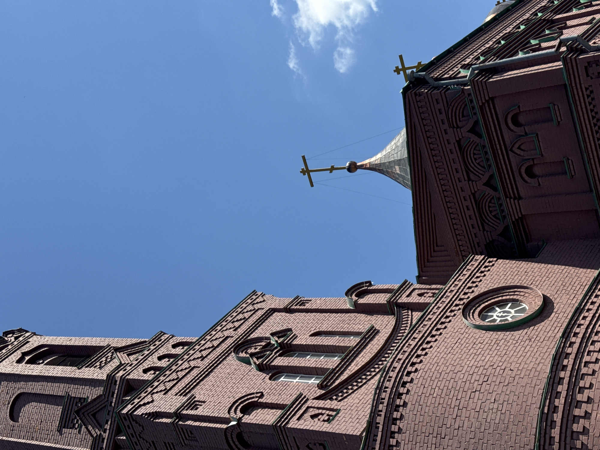 A brick building, possibly a church, is shown from a low-angle perspective against a clear blue sky. The building is constructed of reddish-brown bricks and features architectural details such as arched windows, decorative brickwork, and a rounded window with a distinctive cross-shaped emblem. A conical spire with a cross on top rises from one section of the building. Several other crosses are visible, some on the building's roofline. The sky is mostly clear, with a few wispy white clouds. The image appears to be taken on a sunny day.