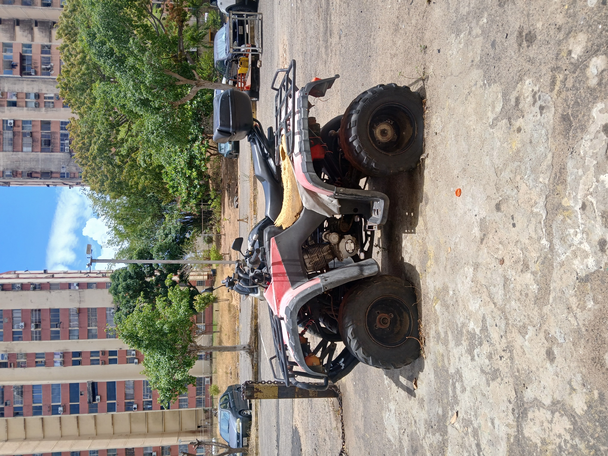 The image shows a dilapidated quad bike parked on a concrete surface in Ciudad Guayana, Venezuela. The quad bike, which is chained to a yellow metal post, appears to be heavily used, with worn tires, faded paint, and general signs of disrepair. The background includes multi-story residential buildings, lush trees, and a partly cloudy sky. A few vehicles are visible in the background as well. The general environment appears urban and residential.
