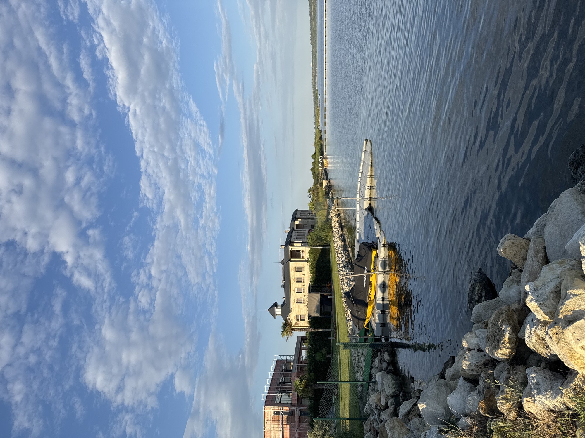 The image captures a serene waterside view in Kazashko, Bulgaria, under a bright sky dotted with white, fluffy clouds. The foreground is dominated by a rocky embankment, with large, light-colored stones and some sparse vegetation. Adjacent to the embankment is a floating dock with a yellow jet ski covered by a black tarp.
To the left, a green fence separates the embankment from a manicured lawn. Beyond the lawn, two buildings are visible. The building on the left appears to be under construction, with scaffolding and exposed brickwork. To its right stands a larger, more elegant structure with a light-colored facade, multiple windows, and a dark, conical roof topping a central tower. Palm trees and lush greenery surround the buildings, creating a sense of a well-developed waterfront property.
The water itself is calm, with gentle ripples reflecting the sky. In the distance, the expansive lake stretches towards the horizon, where a long, thin structure, possibly a pier or a breakwater, is visible. The overall atmosphere is tranquil, suggesting a peaceful day in this Bulgarian lakeside community.