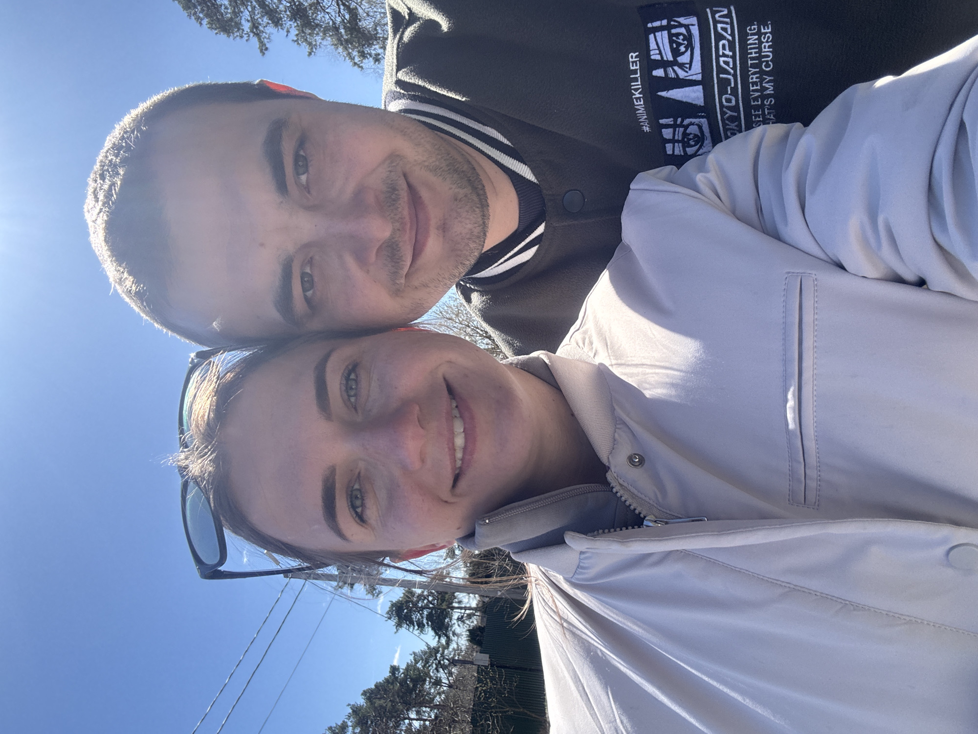 A selfie taken by two people, a man and a woman, outside on a sunny day. The man is on top wearing a dark jacket with white stripes on the collar and sleeves and a graphic that includes the text "ANIMEKILLER TOKYO-JAPAN." The woman is below, wearing a light-colored jacket and sunglasses on her head. The background shows a clear blue sky, power lines, trees, and possibly houses. The location is in Раздолье, Russia.