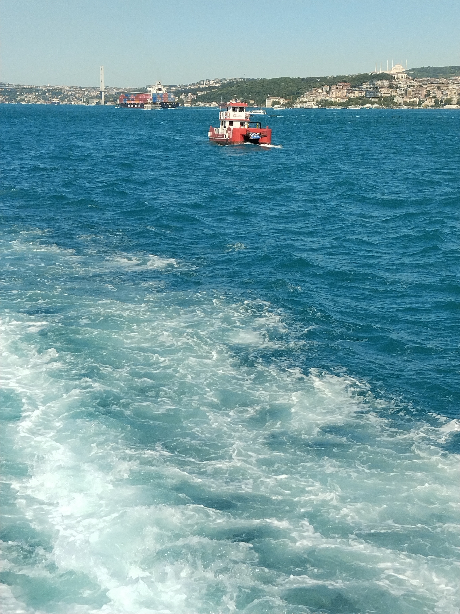 The image captures a view of the Bosphorus Strait, presumably taken from a vessel. In the foreground, the agitated waters show a wake, indicating movement. A red and white boat is positioned mid-ground, moving from right to left. Further in the distance, a large cargo ship is visible, accompanied by the silhouette of the Yavuz Sultan Selim Bridge. The distant shore features a cityscape, likely within Sarıyer, Turkey, characterized by residential buildings and visible landmarks. The scene is bathed in clear daylight, with a blue sky overhead.