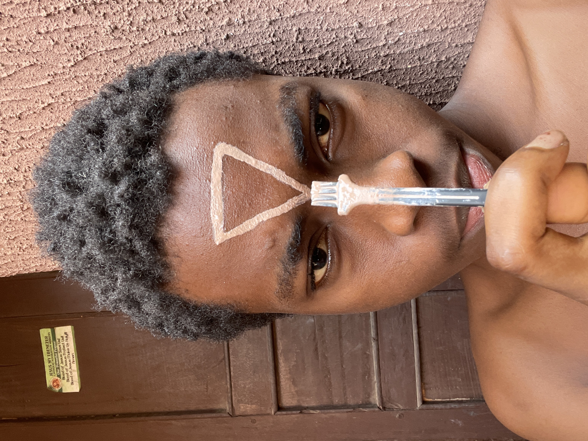 A person with short, dark, curly hair is lying on their side, with a brown textured surface behind their head and a brown wooden door with vertical panels in the foreground. They are painting a light brown triangle on their forehead with a makeup brush. A small sticker with the text "JESUS MY EBENEZER CATHOLIC ADORATION MINISTRY," and the prayer "Blood of Jesus Save Usl Blood of Jesus Protect Usil Blood of Jesus Make Us Holy Amen," is visible on the door. The photo was taken in Nkpor, Nigeria.