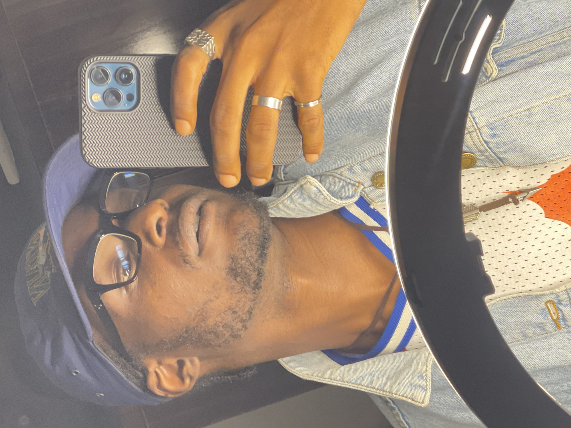 A young Black man with dark skin and short, dark hair is taking a selfie in what appears to be a dimly lit room. He is wearing a dark blue baseball cap, black-rimmed glasses, a light blue denim jacket over a white jersey with orange and blue accents, and several silver rings on his fingers. The background is dark and reflective, suggesting the presence of a mirror or a dark, shiny surface. A ring light is partially visible in the foreground, casting a glow on the man's face and chest. The man is looking at his phone, which he is holding in his right hand. His left hand is resting on his leg. The overall impression is a casual, self-taken portrait. Without further information, it is impossible to determine the specific location in Abuja, Nigeria.