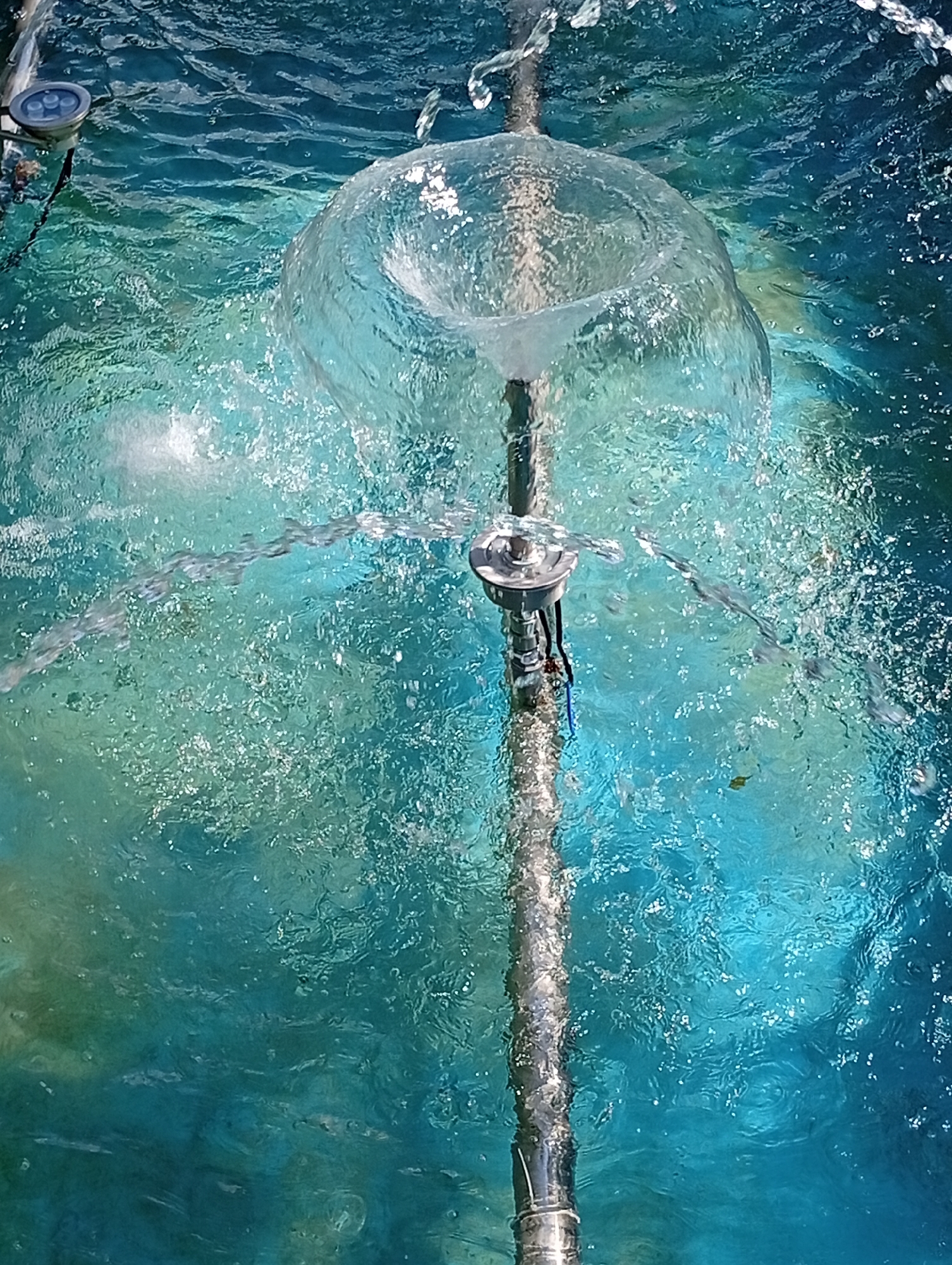 The image shows a close-up view of a water fountain in Kostanay, Kazakhstan. The fountain features a central metal pipe with water spraying outwards in multiple streams, forming a dome-like shape of water at the top. The water is clear and reflects the sunlight, creating sparkling patterns on the surface. Underwater lights are visible in the upper left corner of the frame, emitting a soft blue glow. The water itself is a vibrant turquoise color, suggesting it may be treated or illuminated. The overall impression is one of a refreshing and visually appealing water feature.