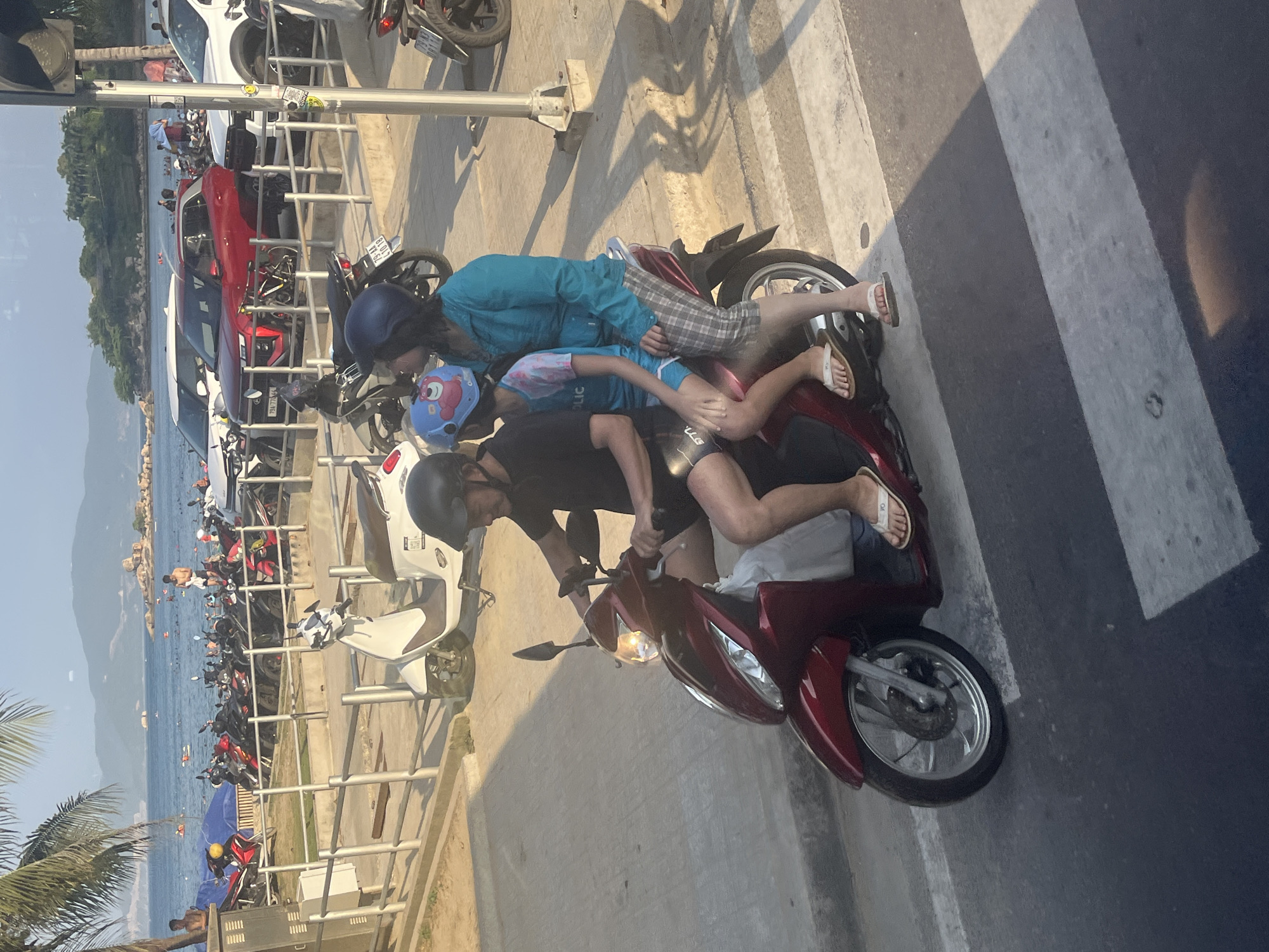 This image captures a family of three riding a red scooter on a street in Nha Trang, Vietnam. The father, wearing a black t-shirt and dark shorts, is driving. Beside him, a child with a light blue shirt and a blue helmet adorned with a cartoon character is seated. Behind the child, a woman in a light blue long-sleeved shirt and plaid shorts is the passenger. They are on a street with a crosswalk, with a line of parked scooters and several cars visible in the background. Beyond the street, a beach area with people swimming and enjoying the water can be seen, with hills and rocky outcrops in the distance. The scene is bathed in the warm light of late afternoon.
