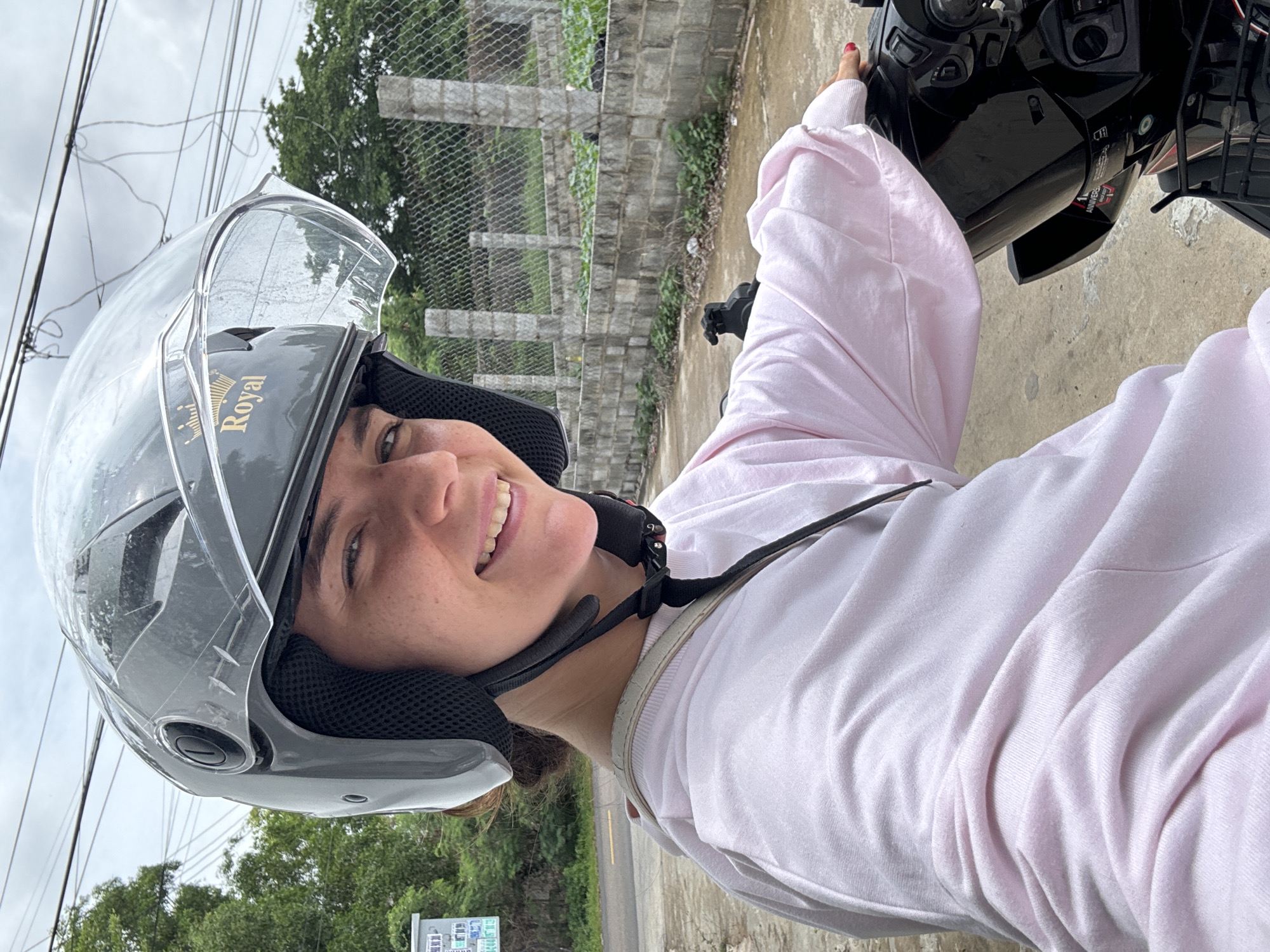 A young woman wearing a gray and clear motorcycle helmet and a light pink hooded sweatshirt is sitting on a black motorcycle. She is looking at the camera and smiling. The helmet has a gold crown logo with the word "Royal" written beneath it. She has her left hand on the handlebars of the motorcycle, and her right hand is resting on the seat. The background shows a gray concrete wall, a chain-link fence, green trees, and some electrical wires overhead. The scene appears to be in an outdoor urban environment, possibly in Mũi Né, Vietnam, given the characteristic architecture and vegetation.