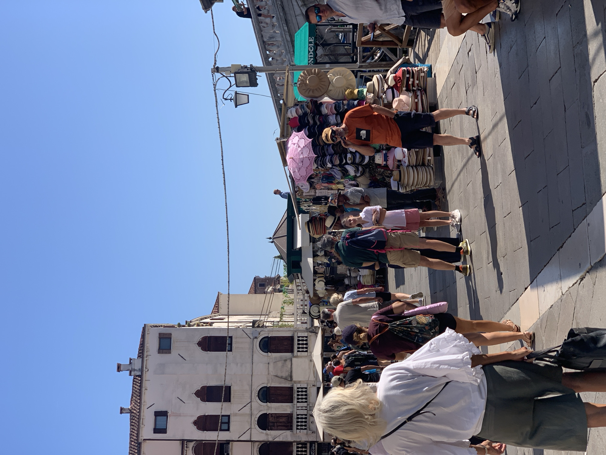 The image depicts a bustling marketplace scene in Venice, Italy, likely on a sunny day. In the foreground, a woman with blonde hair wearing a white ruffled top and olive-green shorts is walking away from the camera, carrying a black tote bag. She is on a paved walkway.
Behind her, a crowd of people are browsing various stalls. One stall is laden with a colorful assortment of hats and an open pink umbrella. Another stall displays more hats, alongside other merchandise like scarves. Men and women in casual summer attire are visible, some examining the goods, others walking through the crowded area.
The architecture in the background features traditional Venetian buildings with tiled roofs, arched windows, and balconies. Power lines are strung overhead. A bridge can be seen in the upper right portion of the image, hinting at the city's canal network. The sky is clear and blue. The overall atmosphere suggests a lively tourist area, common in Venice during peak season.