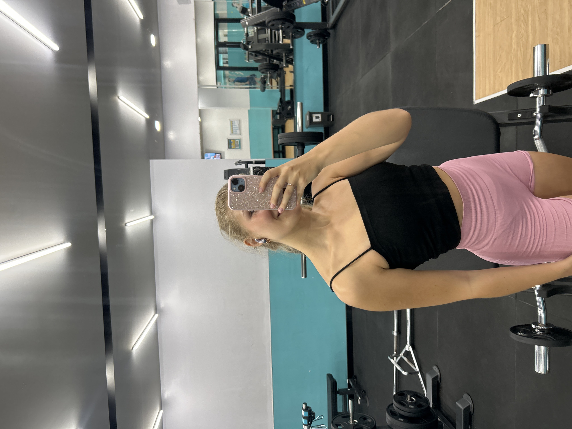 The image depicts a woman taking a selfie in a gym. She is wearing a black tank top and pink shorts. The gym is equipped with weightlifting equipment. The location is inferred to be a gym in Kuta Selatan, Indonesia, based on the user's prompt, but this cannot be definitively confirmed from the image alone.