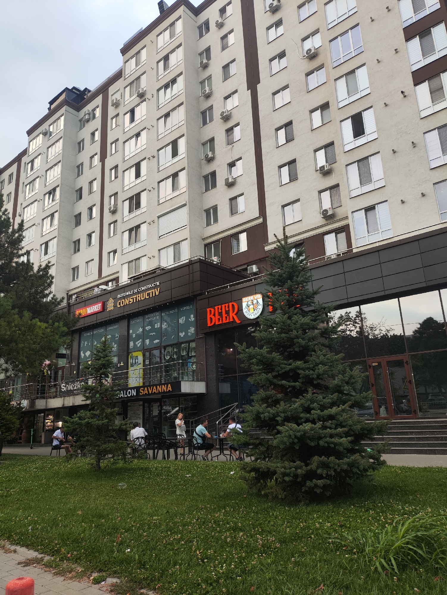 The image shows a multi-story building with commercial establishments on the ground floor, located in Chișinău, Moldova. The building features a light-colored facade with numerous windows and air conditioning units on the upper floors. The ground level has a dark-colored facade with large glass windows and several signs for businesses, including "MATERIA DE CONSTRUCTII CONSTRUCTIV," "Snieżko," "SALON SAVANNA," and a prominent "BEER" sign with a logo. Outside the building, there are tables and chairs set up on a sidewalk, with several people seated. A large evergreen tree stands in front of the building, and a patch of grass with clover is visible in the foreground. The sky is overcast. The context suggests a commercial and residential area within the city.