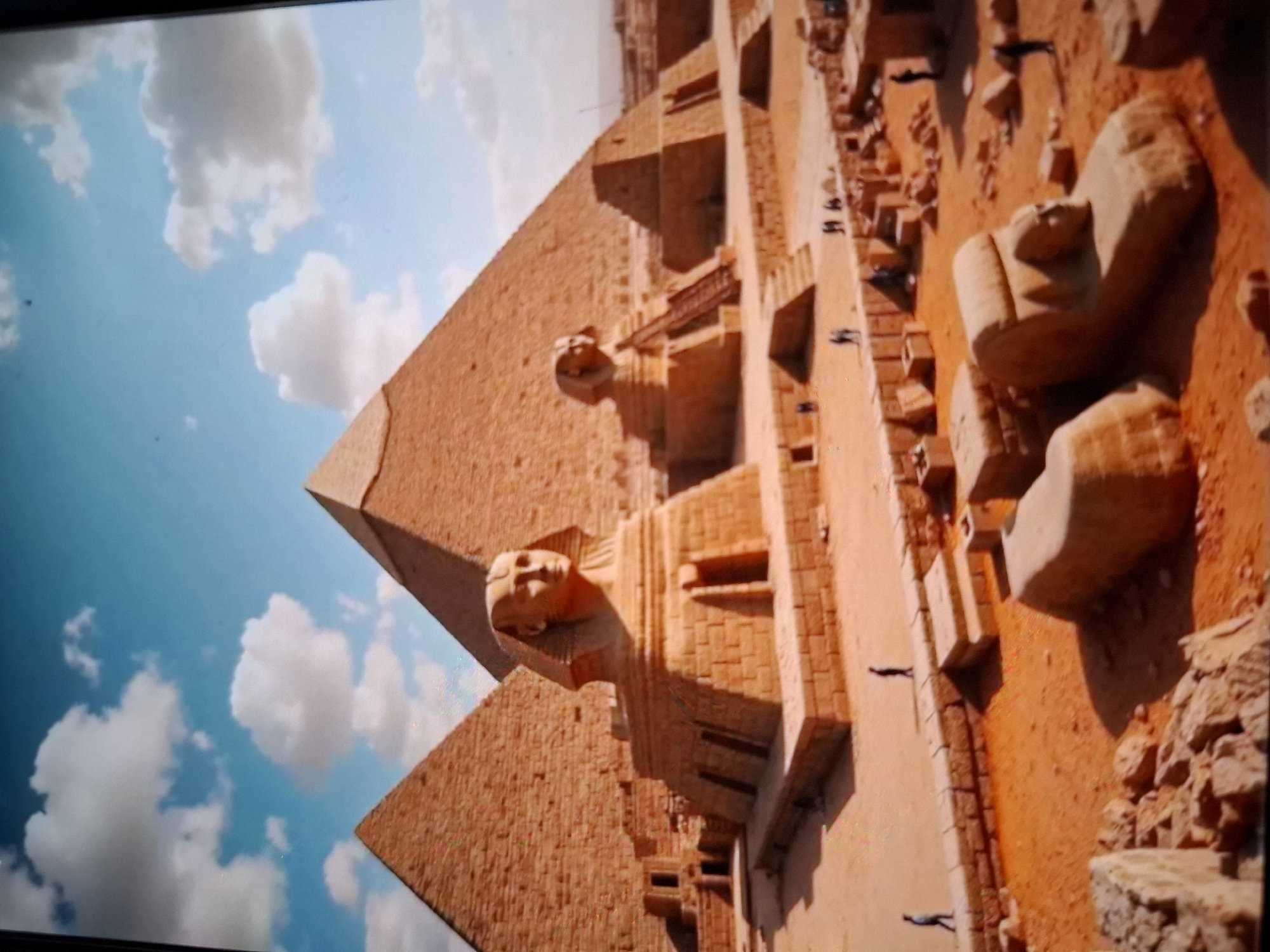 The image depicts the Great Sphinx of Giza and several pyramids under a blue sky with scattered white clouds. The Sphinx is carved from limestone and faces the viewer. It is situated in a sandy, arid environment with other stone structures and blocks scattered around. Several small figures, presumably tourists or workers, are visible in the foreground and midground, walking or standing near the historical monuments. The scene is bathed in sunlight, casting shadows on the sand and structures.