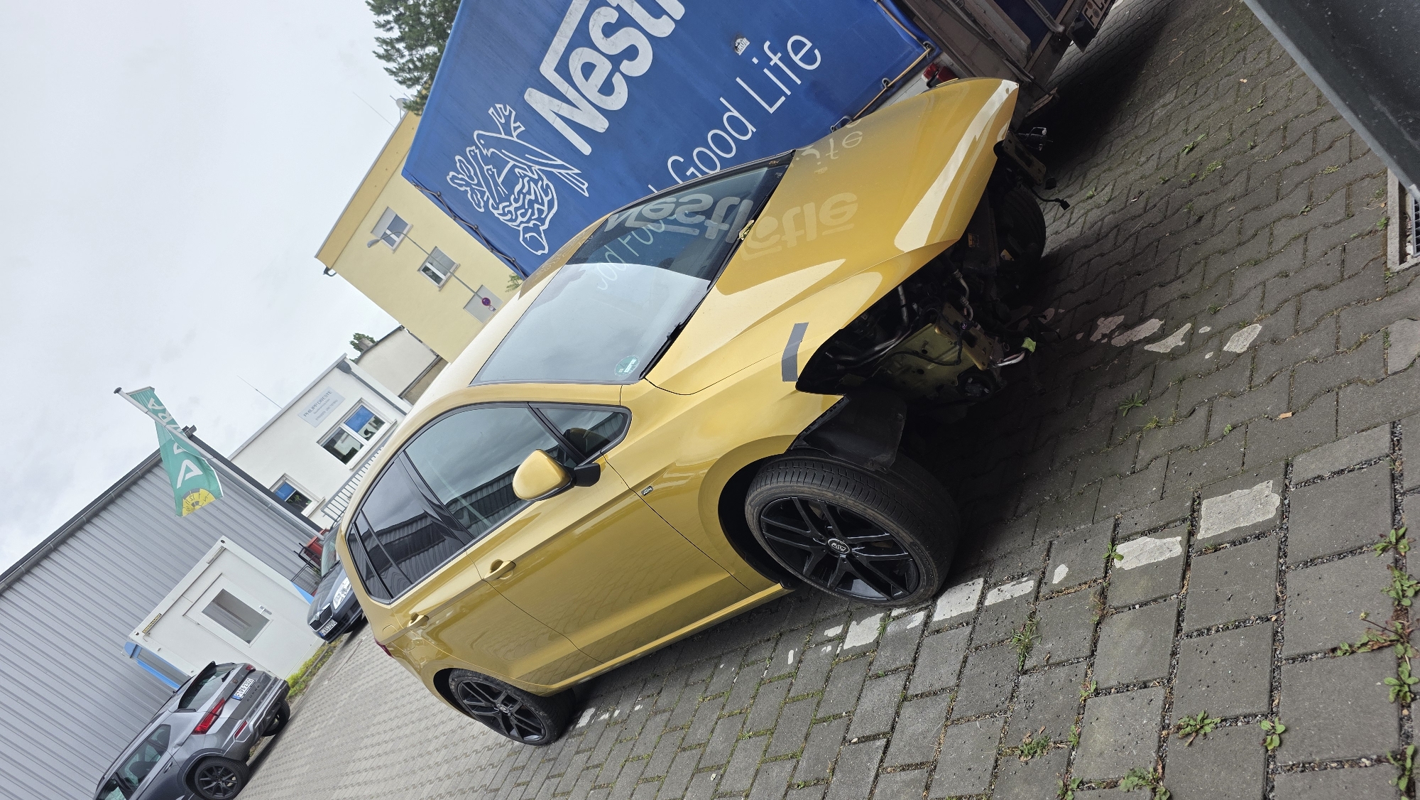 The image shows a yellow car with its front end heavily damaged, parked on a paved surface in what appears to be an outdoor industrial or commercial area. The car is positioned at an angle, with its front bumper and grille missing, exposing the engine components. One of the front wheels is visible, along with a black rim. To the right of the yellow car, a blue truck trailer with the Nestlé logo and the slogan "Good Life" is partially visible. In the background, there are several buildings, including a light-colored industrial building with a grey corrugated metal facade and a yellow building with windows. A flag with a green and yellow design is also visible on a flagpole in the middle ground. A dark gray SUV is parked to the left of the yellow car, and a dark-colored sedan is parked behind it. The ground is covered in rectangular paving stones with some weeds growing in the cracks. The sky is overcast and cloudy. The scene appears to be a street or parking lot in an urban or semi-urban environment, possibly a car repair shop or a logistics hub.