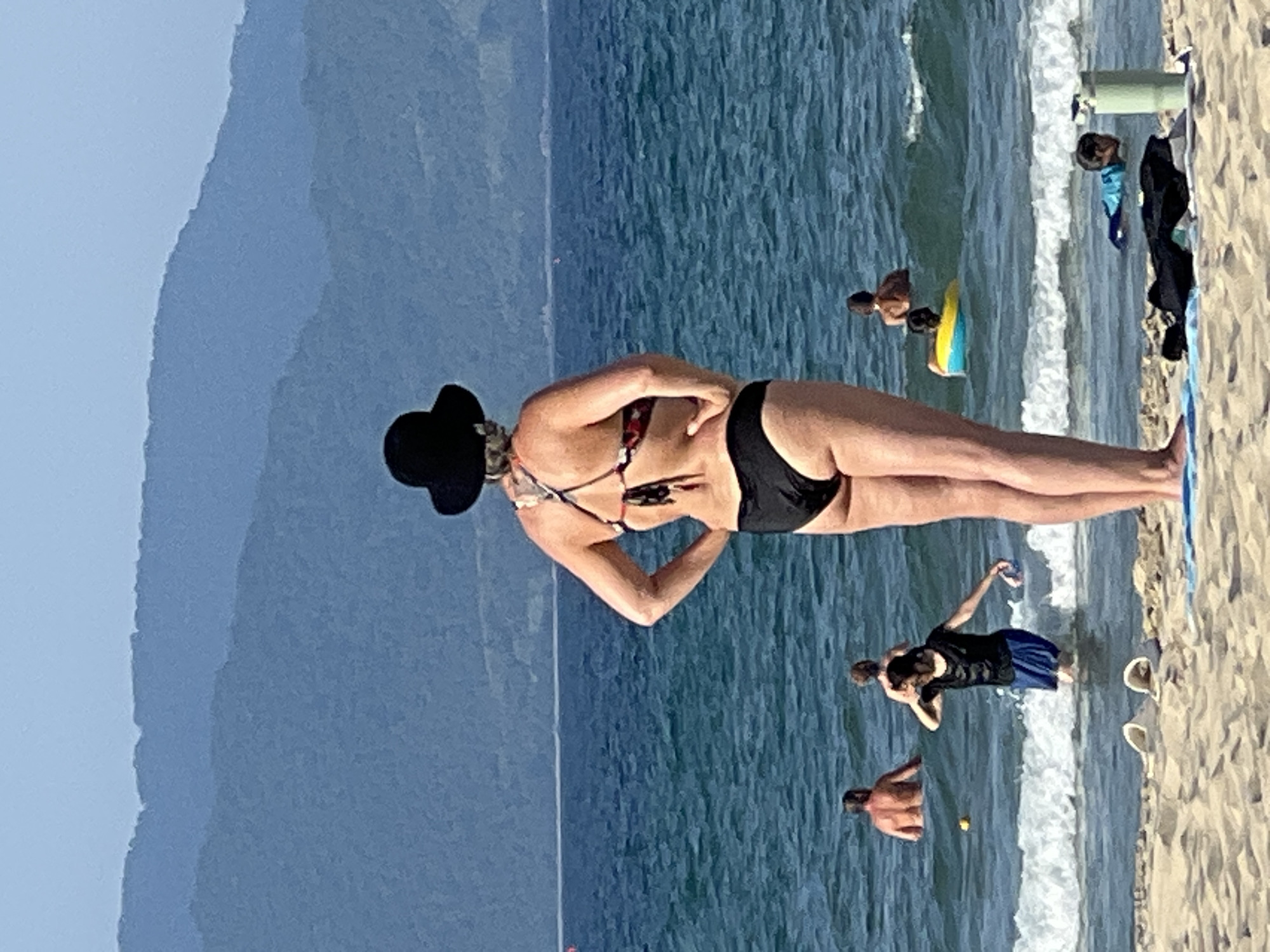 The image depicts a beach scene in Da Nang, Vietnam, with the ocean in the foreground and a hazy mountain range in the background. In the mid-ground, several individuals are visible in the water. Closest to the camera, a woman wearing a black bucket hat and a patterned bikini stands facing away from the viewer, with her hands on her hips. Further out in the water, several people are swimming or wading. A child is visible in the foreground on the beach, near a beach towel and a thermos. The water is blue and moderately choppy with waves breaking near the shore. The sand is light brown.