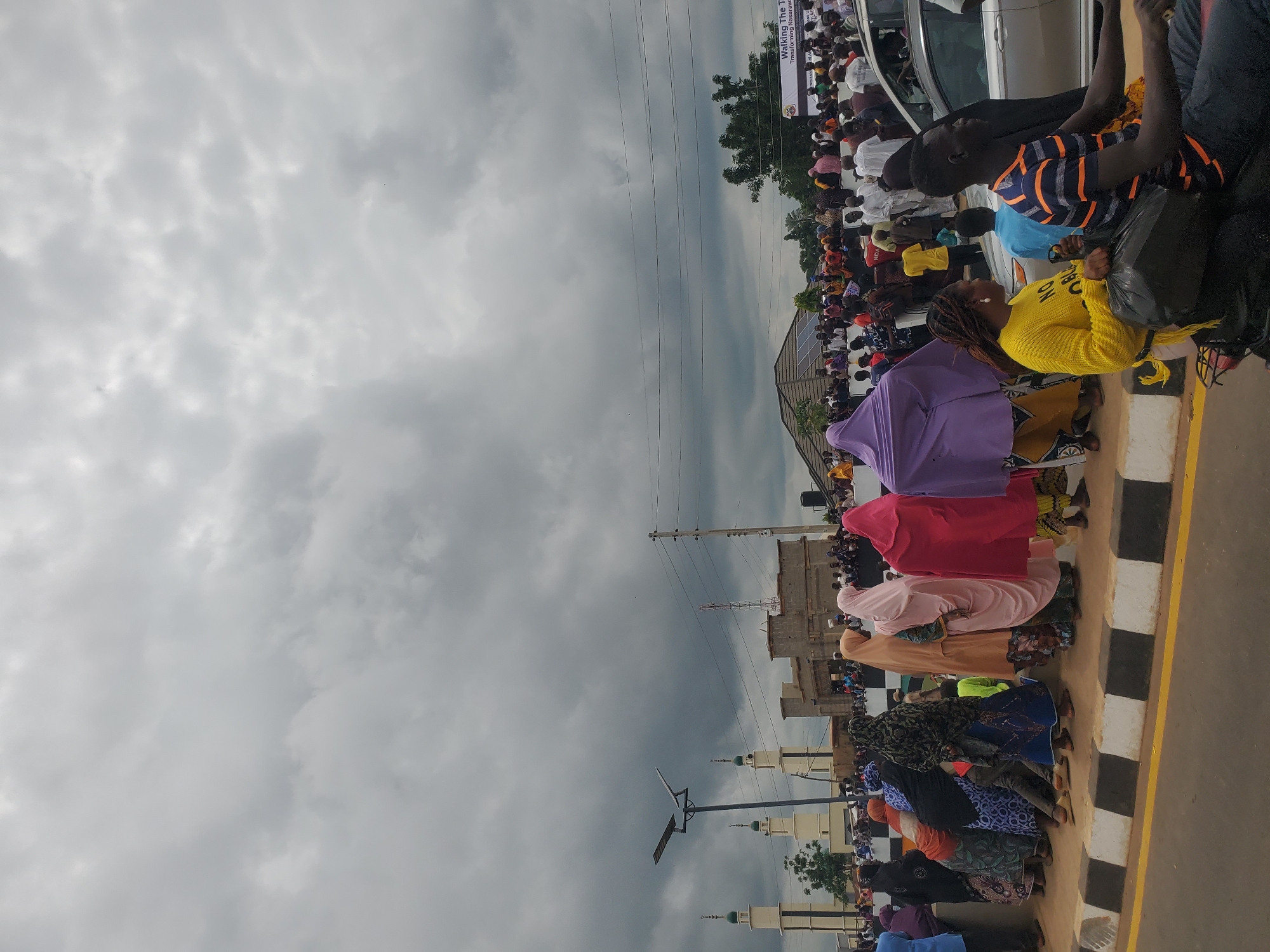 The image shows a street scene in Akwanga, Nigeria. A crowd of people, many wearing traditional clothing, are gathered along a street, possibly for an event. In the background, two prominent mosques with white minarets are visible, along with other buildings and utility poles. The sky is overcast with heavy clouds. A white banner with text, including "Walking The T," is displayed on the right side of the image. People are standing, sitting, and some are on a motorcycle in the foreground. The road has a black and white striped curb and a yellow line.