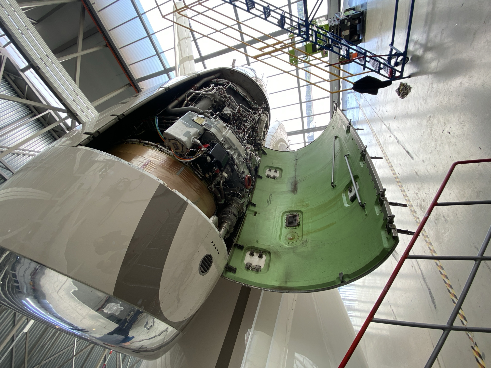 The image shows the open engine bay of an aircraft, likely during maintenance, inside a hangar in Skulte, Latvia. The engine is exposed, revealing its complex internal components. The access door is open, revealing its light green interior surface. A maintenance scaffold is visible above the aircraft. A red and gray ladder stands near the open engine bay. The hangar features a corrugated metal roof with translucent panels providing natural light. The floor is concrete. The location is identified as Skulte, Latvia. There are no people visible in the immediate foreground.