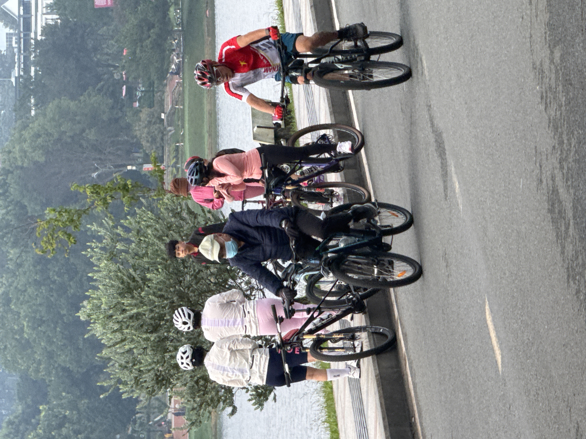 In Da Lat, Vietnam, six individuals on bicycles are seen on a paved road next to a body of water. The group includes two women in white tops and helmets, a woman in a pink top and helmet, a man in a dark jacket, face mask, and helmet, and a man in a red and white cycling jersey, helmet, and sunglasses. The background features lush green trees and a cloudy sky. The scene captures a moment of cycling activity by the lakeside, possibly in Xuan Huong Lake area given the landscape.