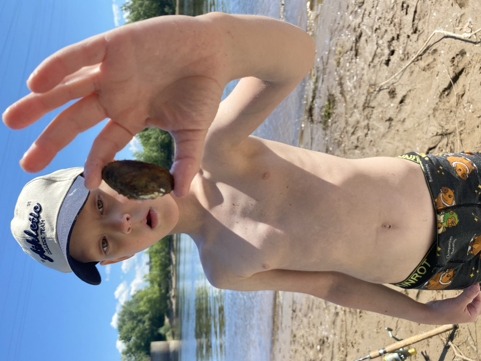 A young boy is holding up a shell to the camera. He is wearing a white baseball cap with blue lettering that reads "COLLEGE TEAM" and has a blue brim. His shorts have a pattern of cartoon animals, possibly chipmunks or groundhogs, and stars. The boy has a surprised expression on his face with his mouth slightly open. He is standing on a sandy bank next to a body of water, likely a river, with trees and a blue sky visible in the background. The sun appears to be shining, suggesting it is a clear day. The overall scene suggests an outdoor activity, possibly fishing or exploring by the water. Fishing rods can be seen on the ground in the lower right portion of the image. The text "UNROT" is visible on the waistband of the boy's shorts. The photo was taken in Chaikovsky, Russia.