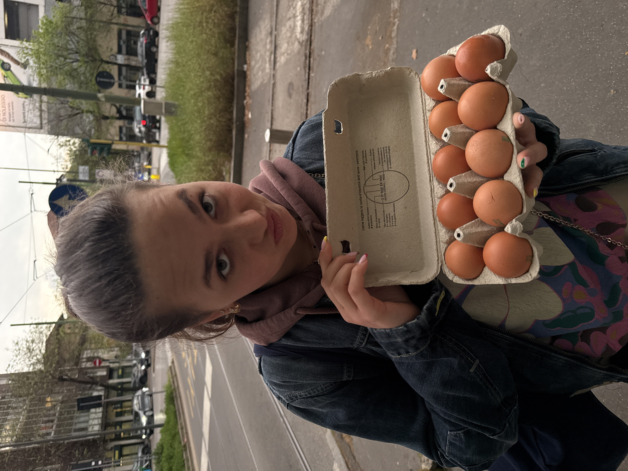 A medium-shot image depicts a young woman holding a carton of brown eggs. She is dressed in a blue denim jacket over a mauve hooded sweatshirt and a patterned top. The eggs are arranged in a typical cardboard egg carton, and some have what seems to be paint on the shells. She is looking directly at the camera with a slight tilt of her head.
The background shows a street scene that suggests an urban setting. Cars are visible on a road, and there's greenery and a building in the background. A street sign is also visible. The location is Milan, Italy.