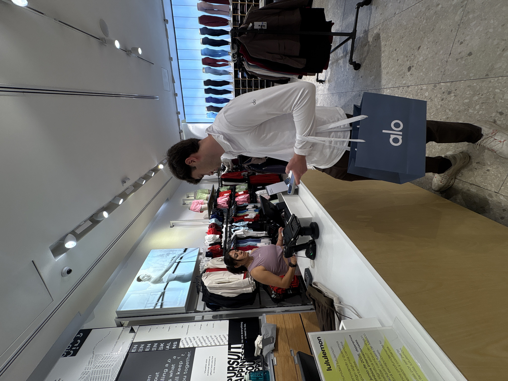 The image depicts an indoor scene within a modern activewear retail store, likely in Puebla, Mexico, given the provided context and visible Spanish text. A man with dark hair, wearing a white long-sleeve shirt with an "alo" logo and dark pants, stands in the foreground. He holds a blue "alo" shopping bag and appears to be looking down at his phone or the counter. He is also wearing white and gray sneakers.
Behind a counter, a woman with dark hair, dressed in a light purple sleeveless top and plaid shorts, is seated. She is looking at a screen or tablet, likely a store employee attending to her duties.
The store's interior features white walls and ceilings with track lighting. Clothing racks filled with various colored activewear, including leggings and tops, are visible in the background. Several digital displays are mounted on the walls, one showing a woman in athletic wear, and others displaying text. A security camera is also visible on the ceiling. The flooring is a light gray, textured surface.
Notable text includes the "alo" logo on the shopping bag and the man's shirt. A sign near the counter clearly shows "lululemon." Other visible English text includes phrases like "PURSUIT," "APPINES," "you are everything u need to be," and "Isabel Reyes Barre Instructor." A Lululemon sign in Spanish discusses "comunidad lululemon" and changes occurring in "junio de 2025," specifically mentioning "México," reinforcing the location. The scene captures a transactional moment in a brightly lit retail environment.
