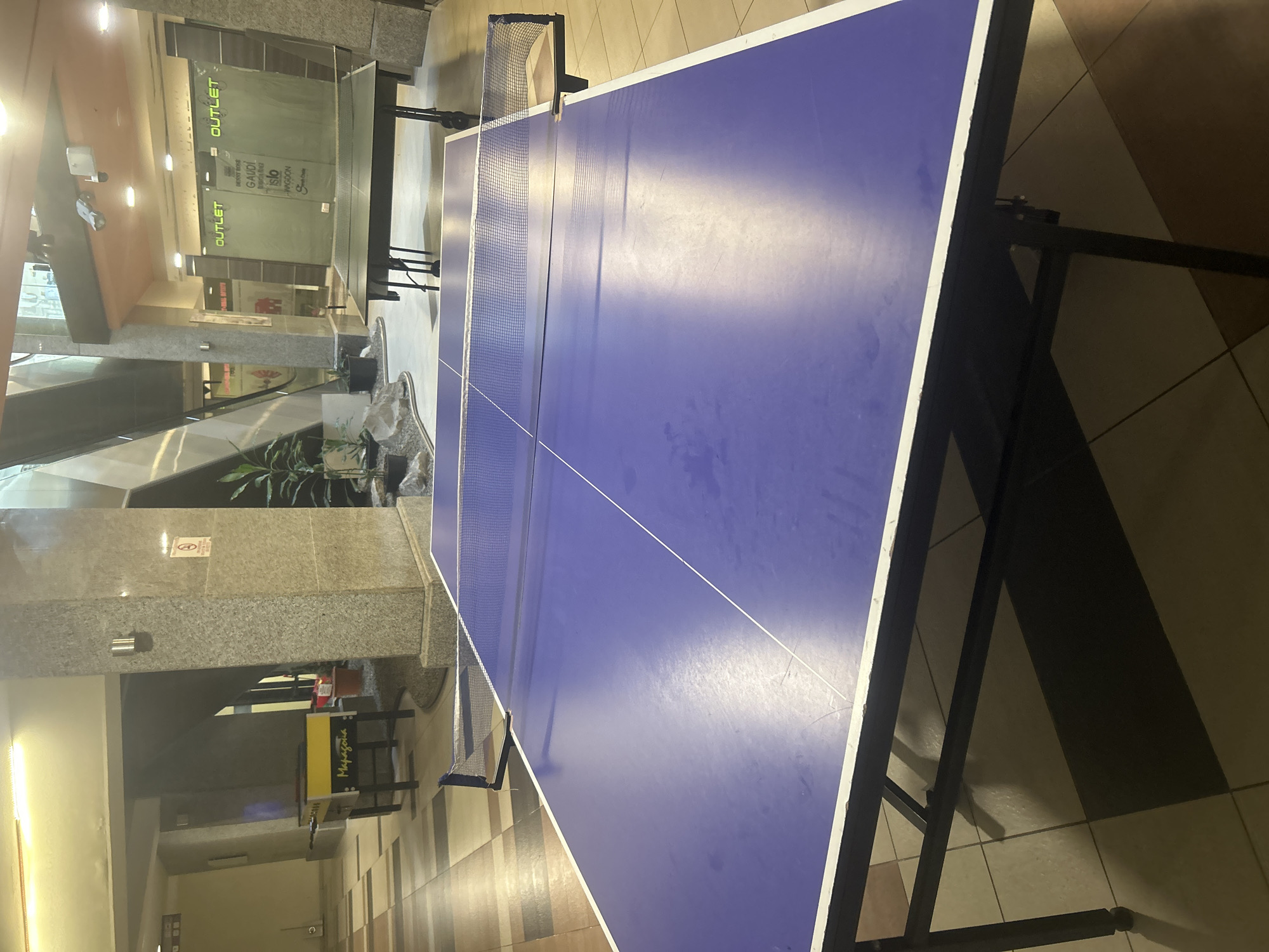 This image captures an indoor scene within a commercial building, likely a shopping mall, in Pleven, Bulgaria. The primary subject is a vibrant blue ping-pong (table tennis) table, centrally located, complete with white playing lines and a net. The table suggests a designated recreational area within the complex.
To the left of the table, several storefronts are visible, two of which prominently display the word "OUTLET" in green lettering. Through their glass windows, various partially legible brand names or shop titles can be seen, including "BOOT BOGS," "GAUDI," "giacomo," "SISLEY," "WAGGON," and "S.Oliver." The interior features grey granite-like pillars and walls, complemented by a tiled floor with patterns in shades of brown and beige.
In the background to the right, an escalator can be seen ascending, indicating a multi-level structure. Further back, a distinctive yellow kiosk or stand is present, bearing the text "Mapagoua." Potted plants are strategically placed, enhancing the environment. A clear "No Smoking" sign, depicted as a red circle with a diagonal line over a cigarette, is mounted on one of the grey pillars.
The space is illuminated by artificial lighting from ceiling and wall fixtures, typical of an indoor commercial setting, making the specific time of day indeterminate. No people are visible, indicating a quiet moment with no discernible activity or event taking place. The scene conveys the atmosphere of a clean, modern, and well-maintained public commercial space.