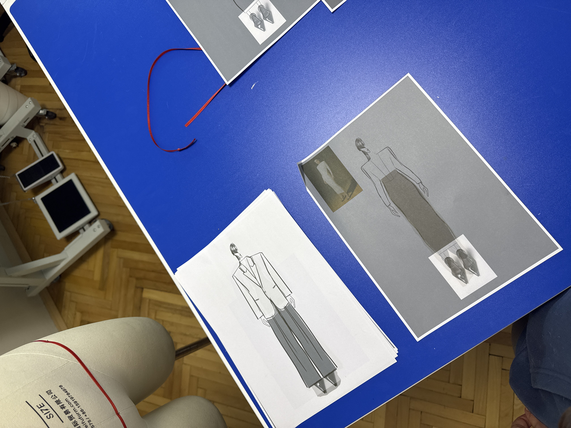 The scene depicts an interior setting, identified as a design studio or workshop in Sofia, Bulgaria. A bright blue tabletop dominates the foreground and midground. On the left side of the table, a stack of white papers presents a fashion illustration of a male figure dressed in a light-colored jacket and dark trousers. To the lower right, a grey sheet features a fashion sketch of a female figure in a long skirt and blouse, with a small photographic image of a woman in a light-colored outfit affixed above it. A second, partially visible grey sheet in the upper right displays an illustration of a pair of pointed-toe shoes. Two distinct pieces of red ribbon lie on the blue table surface in the upper right area.
In the upper left background, a cream-colored dress form, showing the torso and hip sections, is visible. This form features a red line marking a specific point, along with printed text that includes "SIZE," Chinese characters, "meifuform.com," and a phone number. Behind the dress form, white metal studio equipment on wheels supports two dark, rectangular screens. The background flooring consists of light-toned parquet wood arranged in a herringbone pattern. A segment of a person's leg, clad in dark blue fabric, is visible at the extreme bottom left corner of the frame.