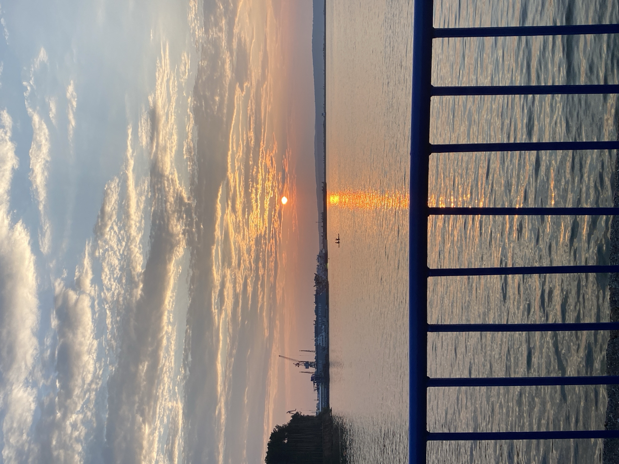 The image depicts a sunset over a wide body of water, likely the Kama River, adjacent to the city of Chaikovsky, Russia. In the foreground, a blue metal railing separates the viewer from the water. The water reflects the warm hues of the setting sun, with a bright orange and yellow path of light stretching across its surface. In the middle ground, a distant shoreline features a line of trees on the left and a harbor with cranes and buildings on the right. Two small figures in a boat are visible on the water. The sky is filled with scattered clouds, tinged with the colors of sunset, ranging from pale blue to warm orange and pink. The overall atmosphere is serene and tranquil.