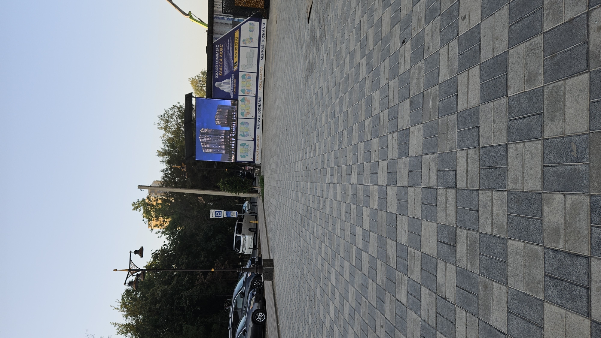 The image shows a street scene in Dushanbe, Tajikistan. In the foreground, a pedestrian walkway is paved with interlocking grey and dark grey rectangular tiles. Beyond the walkway, several cars are parked. To the left, a vintage street lamp with three lights is visible. In the background, there is a large digital billboard displaying an advertisement for a residential complex. The billboard shows an artistic rendering of modern apartment buildings, with text in Russian and what appears to be Persian or Tajik. The text on the billboard includes "ЖИЛОЙ КОМПЛЕКС КЛАССА ЛЮКС" which translates to "LUXURY CLASS RESIDENTIAL COMPLEX," and "MEHVARI DUSHANBE." The environment is outdoors, with trees lining the street and a clear sky overhead. The lighting suggests it is daytime, possibly late afternoon. No people are clearly visible in the image.