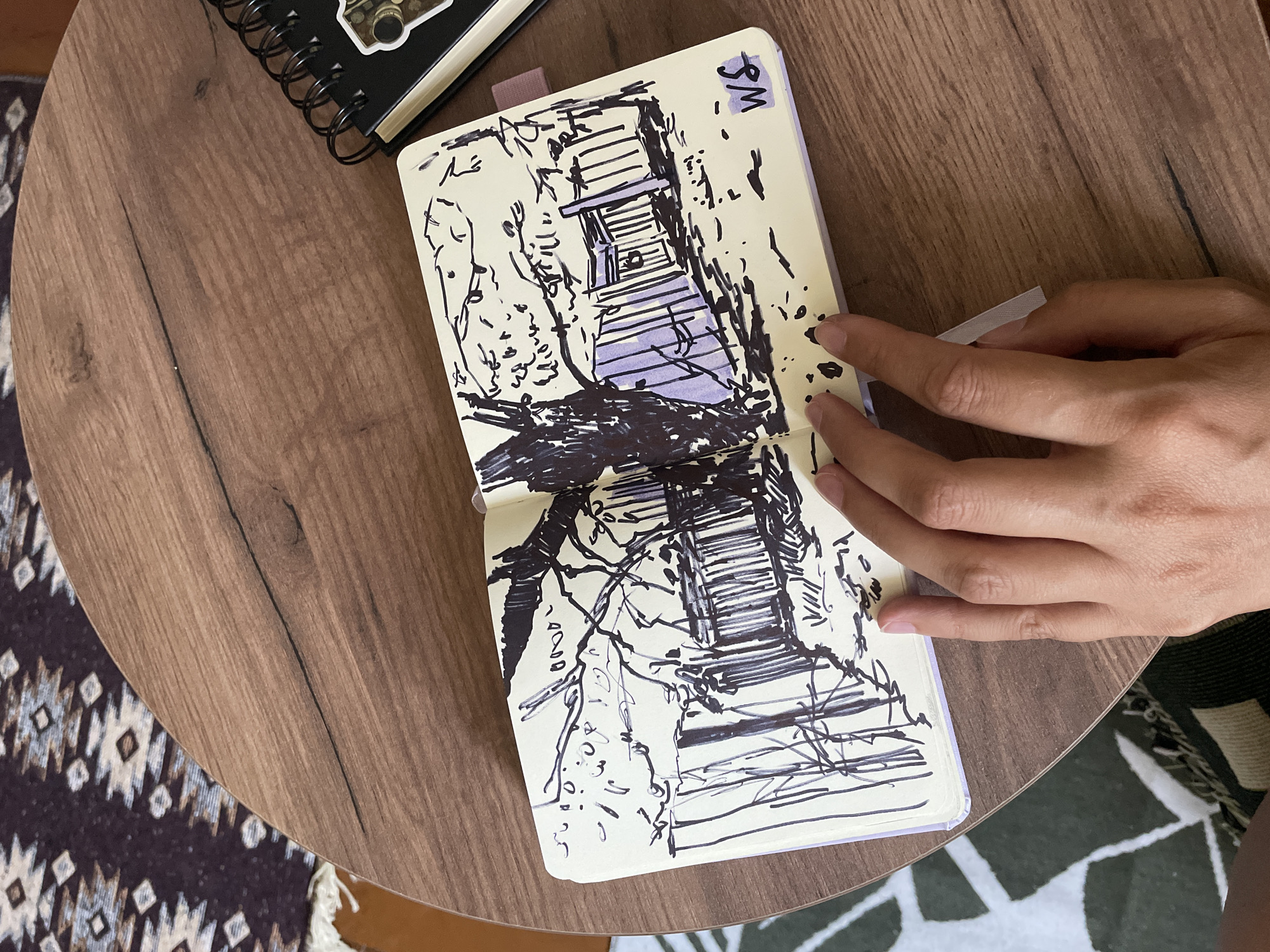 The image shows a top-down view of an open sketchbook with a hand resting on the right page. The sketchbook is open to a black and white sketch of what appears to be a wooden structure, possibly a bridge or a staircase, surrounded by sketched trees and foliage. Some areas of the sketch are filled with purple marker. The sketch is in a notebook with a light-colored cover and a purple tab. The notebook is placed on a round wooden table. To the left of the sketchbook, a black spiral-bound notebook is partially visible. A hand, appearing to be a person's right hand, is gently pressing on the right page of the open sketchbook, with its index finger extended. The background shows a patterned rug beneath the table. The lighting suggests it might be daytime, but no specific time or weather conditions are evident. The word "SM" is written in purple at the top right corner of the sketchbook page.