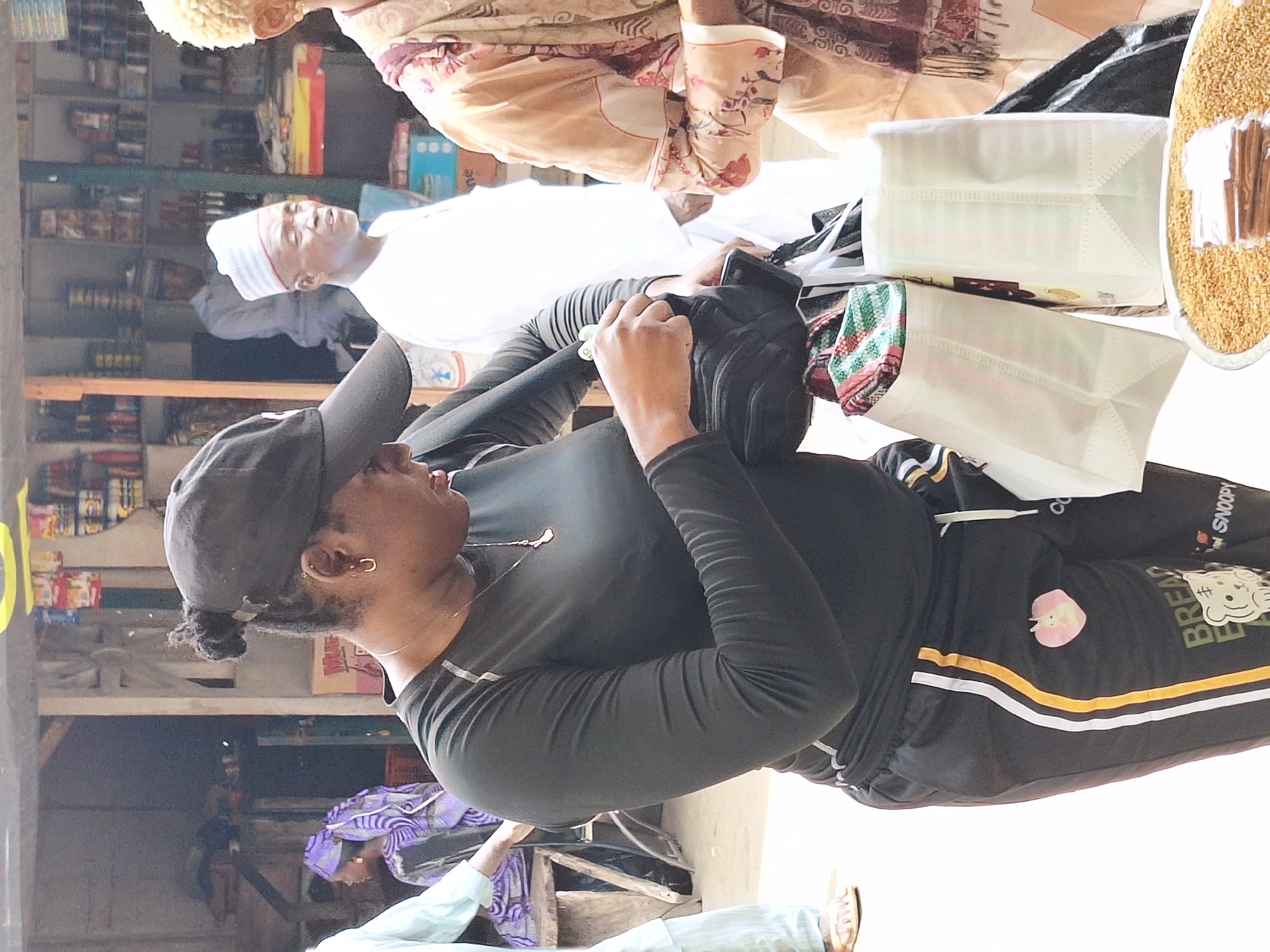 This image captures a bustling indoor market or shop scene in Maiburiji, Nigeria, during the daytime. The lighting is bright, suggesting natural daylight.
In the immediate foreground, a person is visible from the chest up, facing left and slightly upwards. They are wearing a black long-sleeved shirt and black track pants with distinctive yellow and white stripes along the side. A black baseball cap covers their head, and some hair (possibly dreadlocks or braids) is visible beneath it. They wear a gold hoop earring and a thin gold chain necklace. Their left hand holds a black object, possibly a phone or a wallet.
To their left, an older man stands, wearing a white kaftan-style garment and a white cap with red stripes (kufi). He is looking upwards. Further in the background, another person, partially obscured, wears a light-colored, patterned garment.
To the right of the older man, two white non-woven shopping bags are visible on a surface, one containing a green, red, and white patterned fabric. Next to them is an open container filled with light-colored grains or seeds, topped with what appear to be cinnamon sticks.
In the far background, shelves stocked with various products, including bottles and packaged goods, line the walls, characteristic of a local general store or market stall. Parts of another person in light blue clothing and sandals are visible at the bottom left.
Visible text on the track pants of the person in the foreground includes "SNOOPY" and "BREAD EAT", accompanied by a cartoon character and a pink heart-shaped patch. The overall scene depicts a casual interaction or shopping activity within the lively market environment.