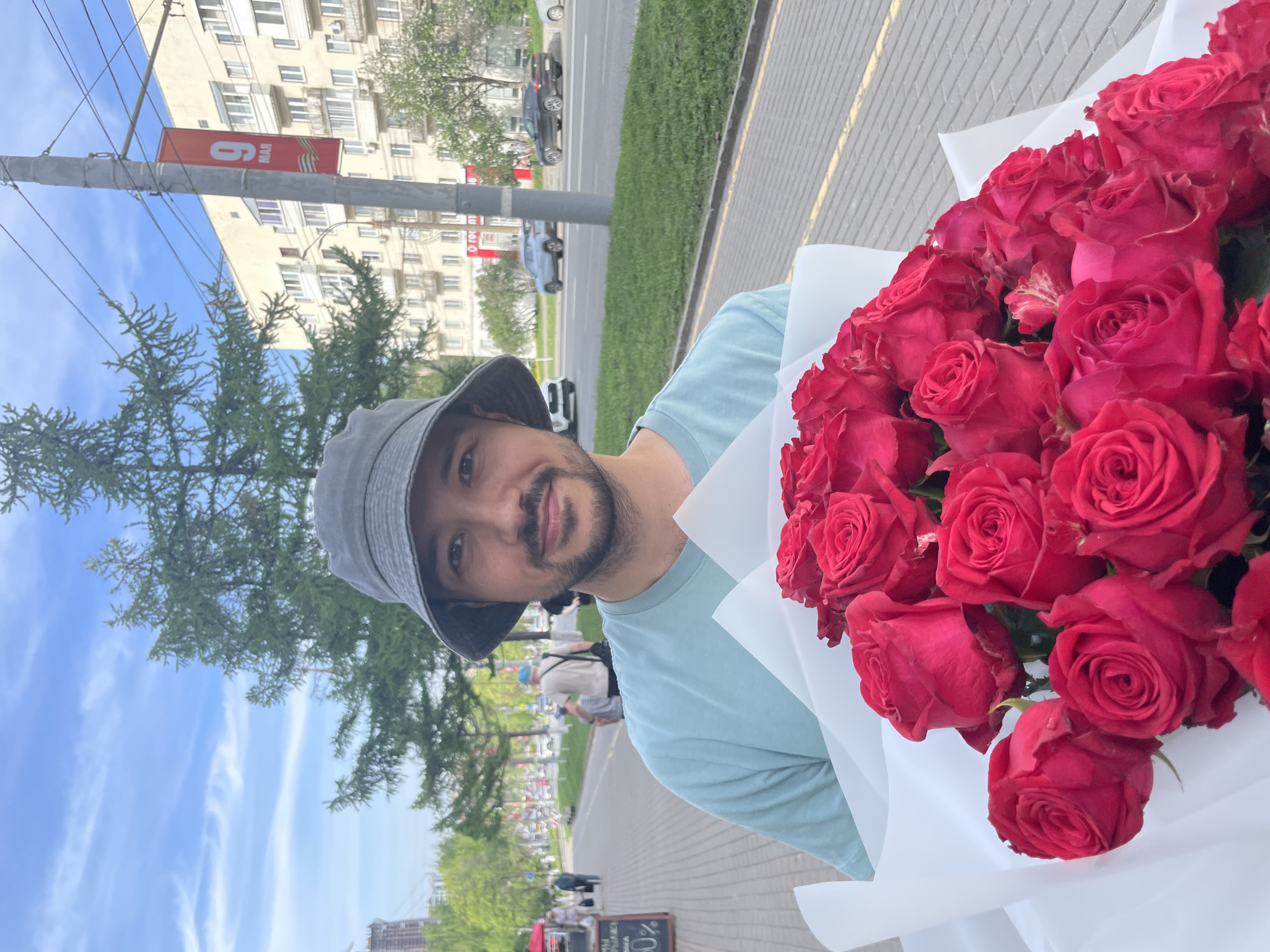 A man holds a bouquet of red roses, likely in Izhevsk, Russia. He wears a light blue t-shirt and a gray bucket hat. A utility pole with a banner displaying "6 MAY" is visible. A building is in the background. There's also a tree, a car driving down a road, and other pedestrians on the sidewalk.