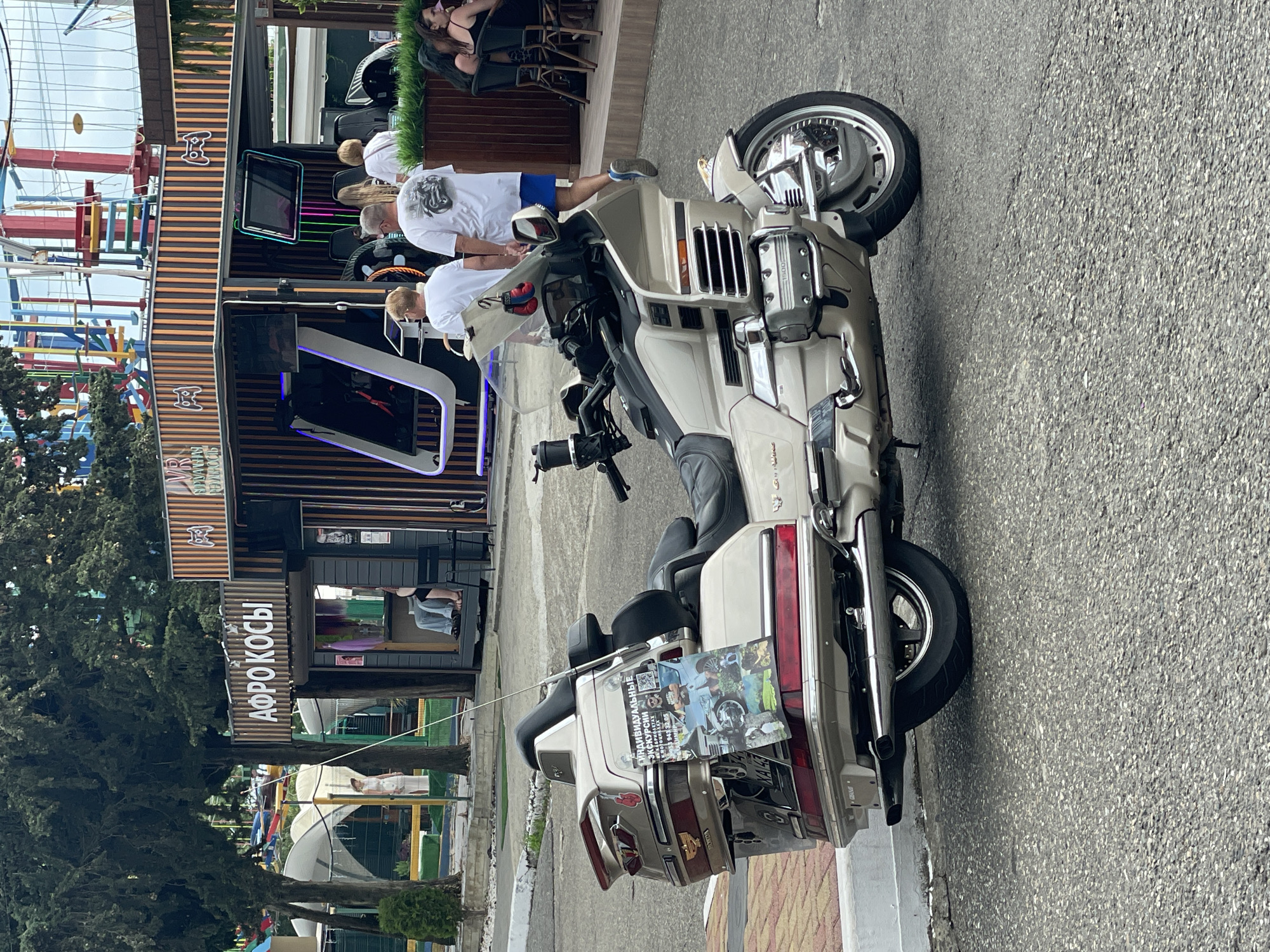 A beige and chrome Honda Gold Wing motorcycle is parked on an asphalt surface next to a curb. A flyer advertising "Individual Excursions" is attached to the motorcycle's rear luggage compartment. In the background, a building with a sign that reads "АФРО КОСЫ" is visible, along with an amusement park attraction and several people. The environment appears to be an outdoor recreational area, possibly a boardwalk or tourist spot.