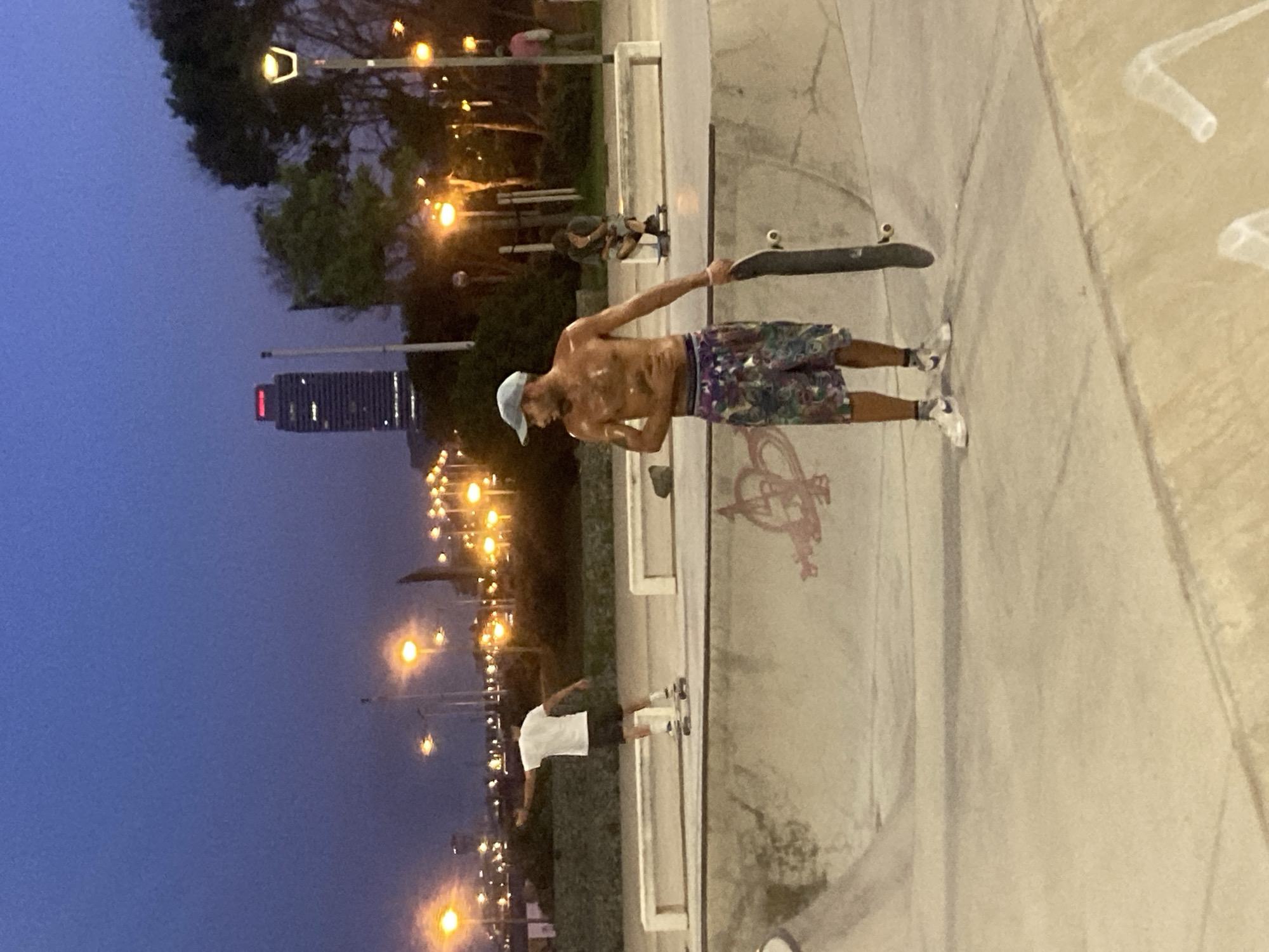 The image depicts a twilight scene at a skatepark, likely in the vicinity of the Port Olímpic area of Barcelona, Spain, given the presence of a prominent, illuminated skyscraper in the background, which resembles the Torre Mapfre or Hotel Arts, and the general urban lighting characteristic of that waterfront district. In the foreground, a shirtless man wearing a light blue baseball cap, patterned shorts, white socks, and athletic shoes stands holding a skateboard in his right hand. His skin appears glistening, suggesting he has been active. To the left and further back in the park, another person on a skateboard is visible, wearing a white t-shirt and dark shorts, their back to the camera. In the midground, a concrete bench is visible, and another person is seated on a skateboard, leaning against the bench. The skatepark itself is constructed of concrete with various ramps and transitions, marked with graffiti. The background is illuminated by numerous streetlights and the lights of the city, with trees and other urban structures contributing to the background composition. The sky is a deep blue, indicative of the evening hours.