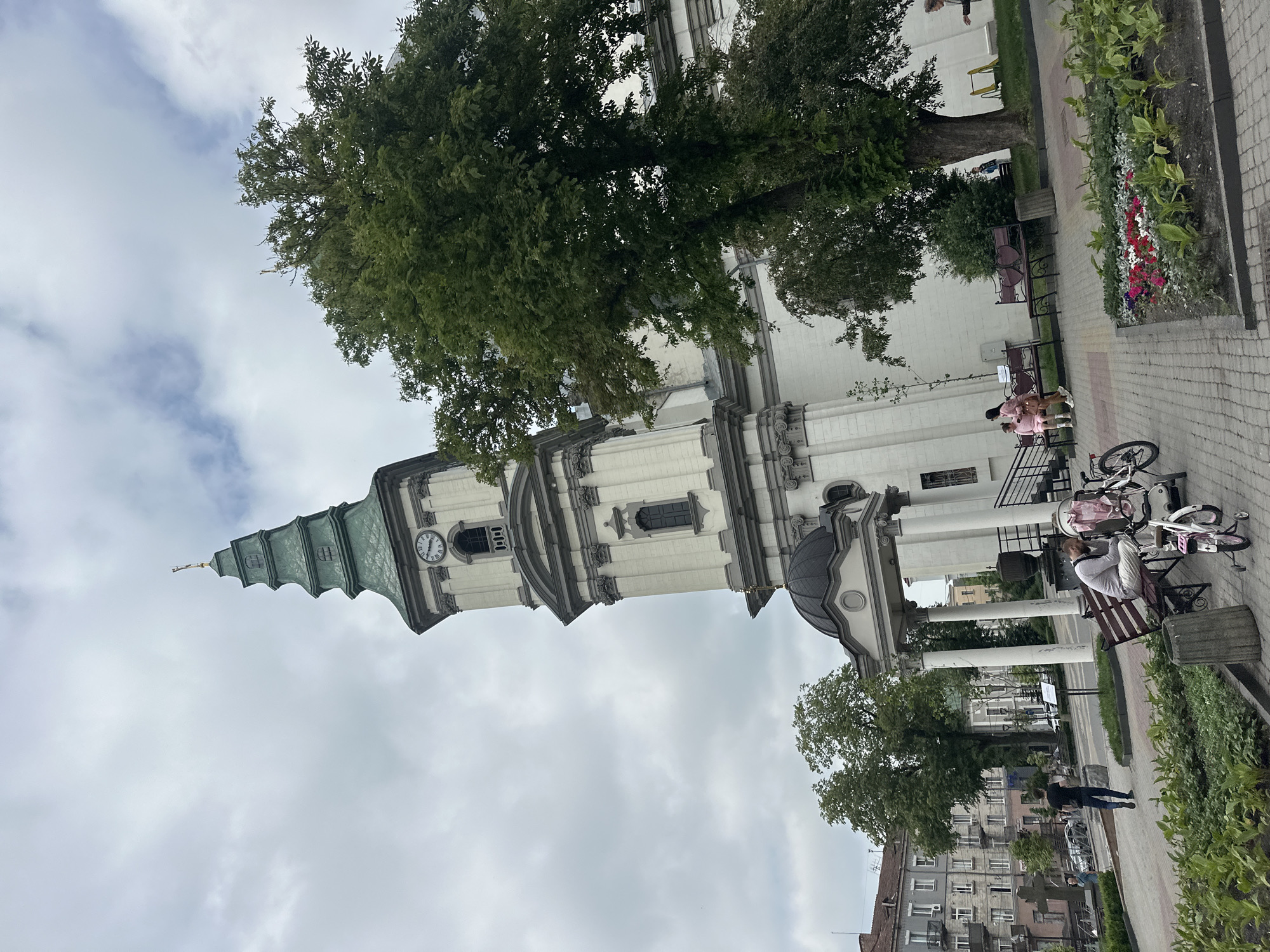 This image depicts the Cathedral of the Immaculate Conception of the Blessed Virgin Mary in Ternopil, Ukraine, on an overcast day.
The central feature is a large, white neoclassical cathedral with a prominent bell tower on its left side. The tower features gray architectural details, arched windows, and a clock face with Roman numerals showing approximately 10:10. The spire of the tower is multi-tiered, green, and topped with a small golden cross. The main entrance of the cathedral is marked by a portico with a dark domed roof supported by white columns. To the right of the cathedral, a large, mature deciduous tree with dense green foliage partially obscures the building.
In the foreground, a paved pedestrian area with gray and reddish paving stones extends across the scene. Several wooden park benches are present. On a bench in the lower center-right, a man in a light-colored long-sleeved shirt and white pants is seated, leaning forward. A pink-lined baby stroller is positioned next to him, along with two small children's bicycles, one pink and one white/pink with training wheels. Further to the right, two women in pink attire are seated on another wooden bench, which features heart-shaped backrests. Flower beds with red, white, and pink flowers, interspersed with green foliage, line the paved areas. Gray concrete trash receptacles are also visible.
In the mid-ground on the left, a man in a dark t-shirt and blue jeans walks away from the viewer on the paved path. Beyond him, several multi-story residential buildings with balconies, primarily in muted gray and brown tones, line a street where parked cars are visible. A large, weathered stone cross monument stands on the far left. The sky is largely overcast with varying shades of gray clouds.