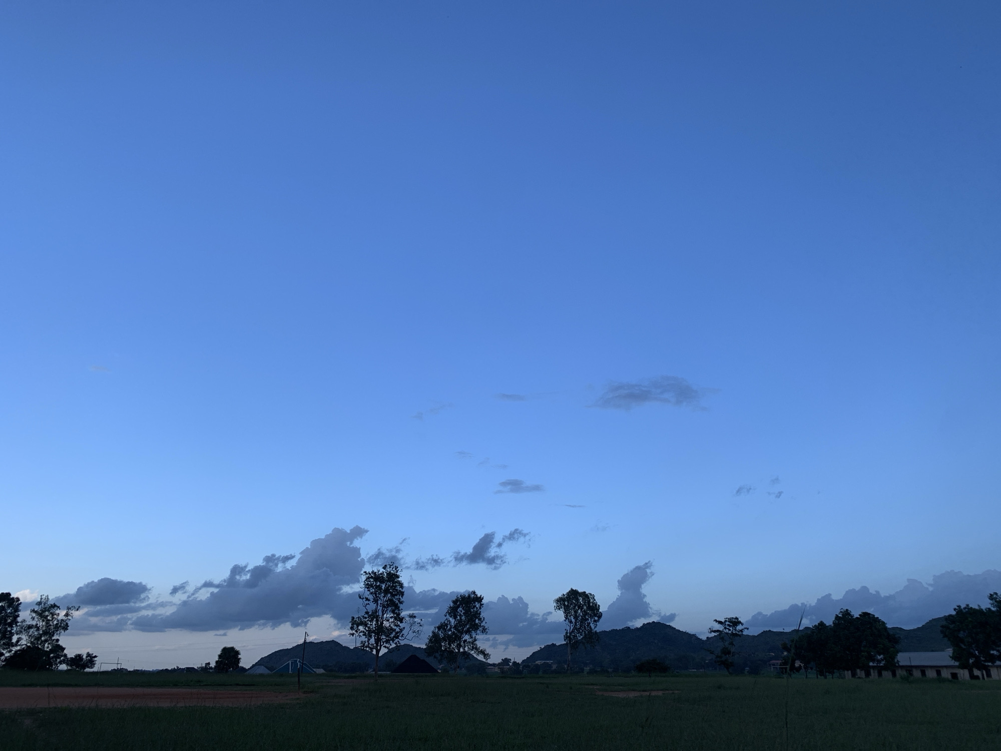 This is a horizontal, eye-level, medium shot of a wide, grassy field under a pale blue twilight sky. The field is mostly dark green, indicating it is likely a lawn or pasture. In the middle ground, silhouetted against the sky, are several dark green trees of varying heights and shapes, and a few dark grey clouds. Behind the trees, a range of dark green, rolling hills stretches across the horizon. A few buildings are visible on the far right, also in silhouette. The image depicts an outdoor scene in what appears to be a rural or semi-rural area. The location in Kuru Karama, Nigeria, is not explicitly visible in the image itself but is provided as context.