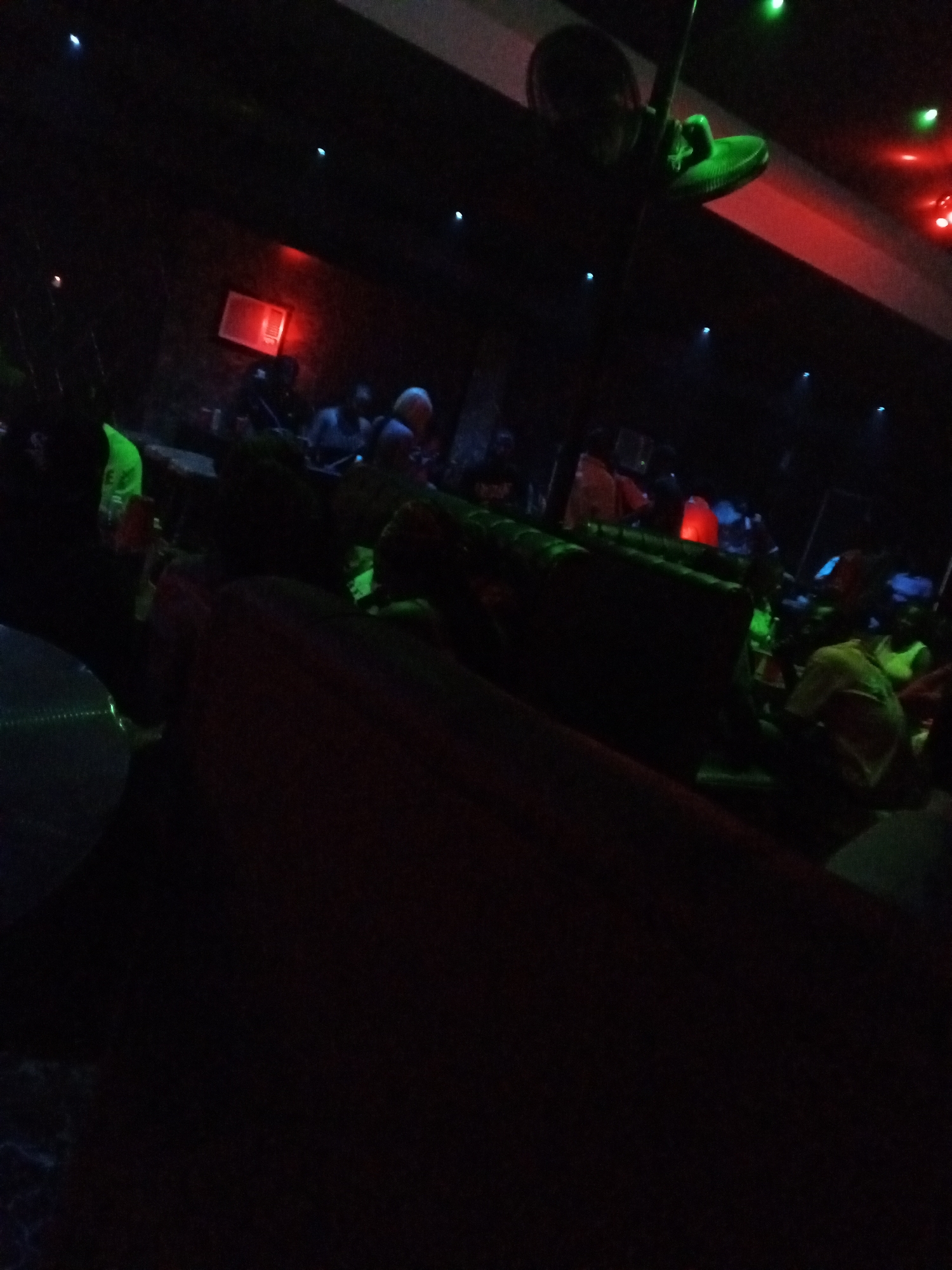 The image depicts a dimly lit interior, possibly a nightclub or lounge in Lokoja, Nigeria. The scene includes several people seated at tables, partially obscured by shadows. A decorative object, possibly a shoe, hangs from the ceiling near a red light fixture. There are dark sofas in the foreground. The overall environment is dark, with strategically placed colored lights providing minimal illumination.