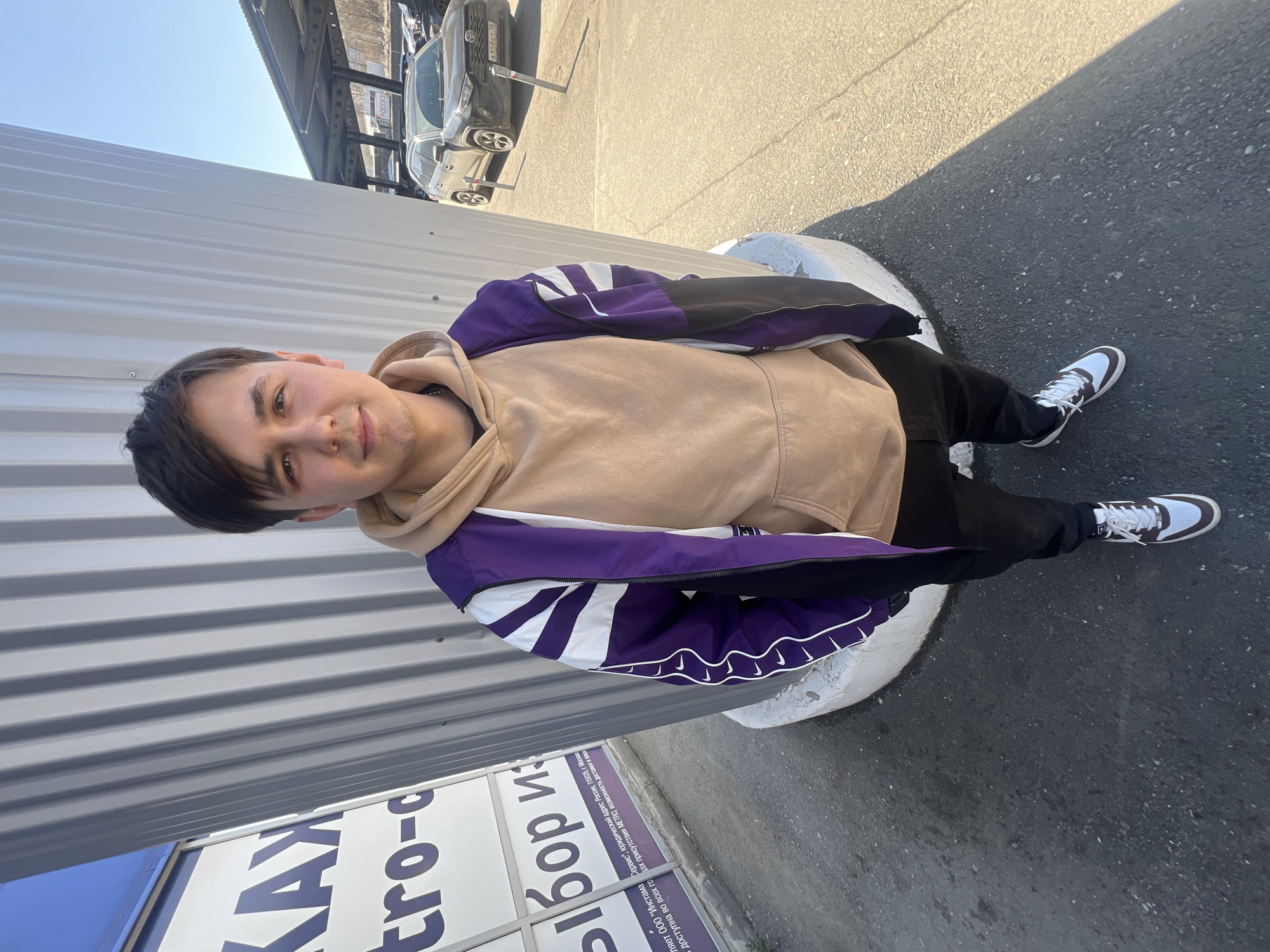 A young man in a beige hoodie and purple jacket stands next to a gray corrugated metal wall with the words "KAK tro-c" printed on a blue and white sign. He is also wearing black pants and white and brown sneakers. He is standing on a white circular base. Behind him, cars are parked under a structure, and the street appears to be paved. In the lower right, the license plate reads "A 958 ET 18", indicating that the location is in Izhevsk, Russia. A segment of text in Russian indicates that the image is in Izhevsk, Russia.