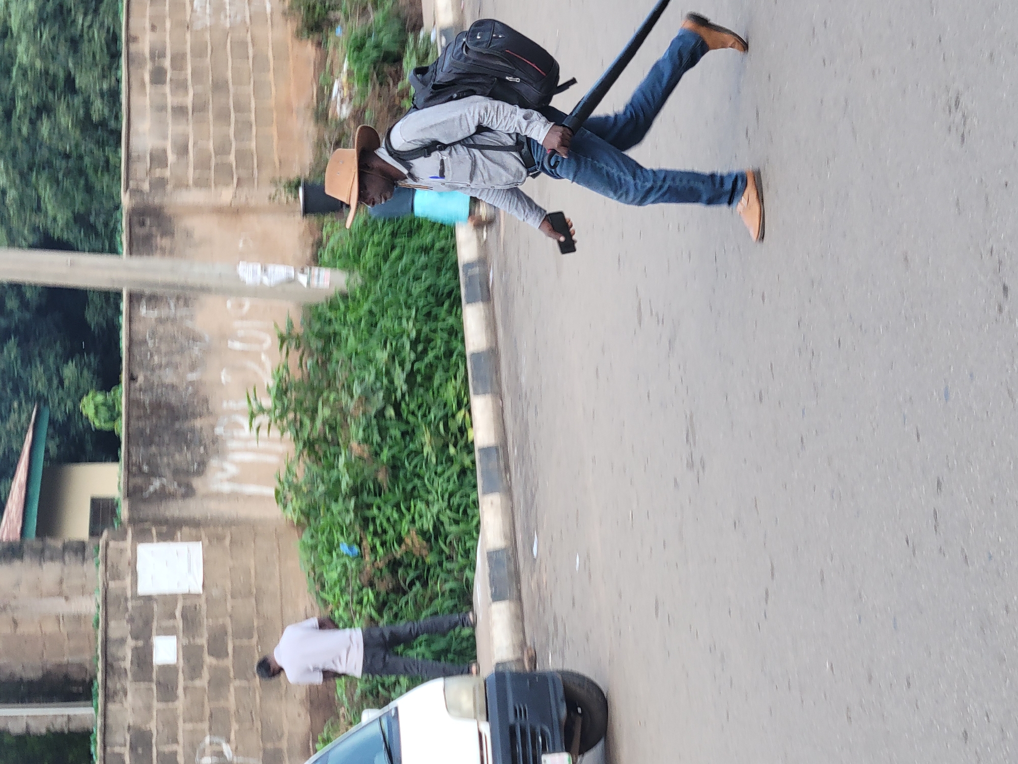 The image depicts a street scene in Barakin Lerawi, Nigeria. In the foreground, a man wearing a light blue long-sleeved shirt, blue jeans, and a brown cowboy hat walks across the road. He carries a large black backpack and holds a black mobile phone in his left hand and a long black object, possibly a tool or equipment, in his right hand. He is wearing brown shoes.
In the background, to the left, a man in a light-colored t-shirt and dark pants is standing near some dense green foliage, facing away from the camera. A white van is partially visible on the far left of the image. Behind the people and foliage, there is a low brick wall and a larger stone wall with some graffiti and posters. Trees and foliage form the backdrop of the scene. The ground is paved with asphalt. The lighting suggests it is daytime.