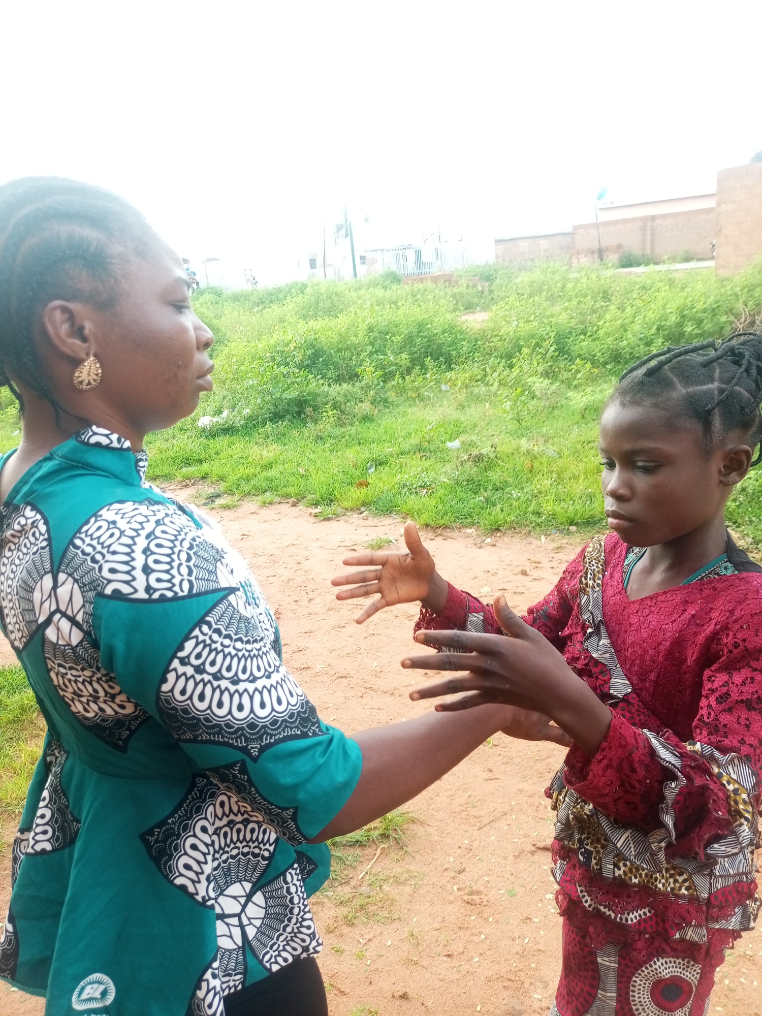 In Tempure, Nigeria, two individuals are captured in an outdoor interaction. An adult woman, from the shoulders up, is facing right, wearing a vibrant teal and white patterned top with a high neckline and intricately braided hair pulled back. A young girl, facing left, is also visible from the shoulders down, dressed in a maroon lace dress with ruffled sleeves and a patterned skirt. She is looking down at her hands, which are positioned forward, as if interacting with the adult woman. The background consists of dry, sandy ground and a patch of green vegetation. In the distance, a light brown brick building with a few vertical white structures is discernible under a bright, overcast sky.