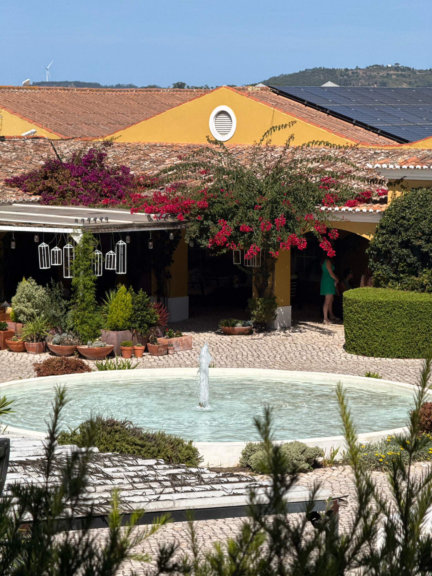This is a wide shot of an outdoor patio area in Mafra, Portugal, with a bright yellow building with terracotta tile roofing in the background. A circular fountain with a central water spout is in the foreground. Several potted plants and trees are arranged around the patio. White geometric lanterns hang from a covered walkway. A woman in a green dress is visible in the background, standing under the walkway. A hedge runs along the right side of the image. Solar panels and a wind turbine are visible on the distant hills.