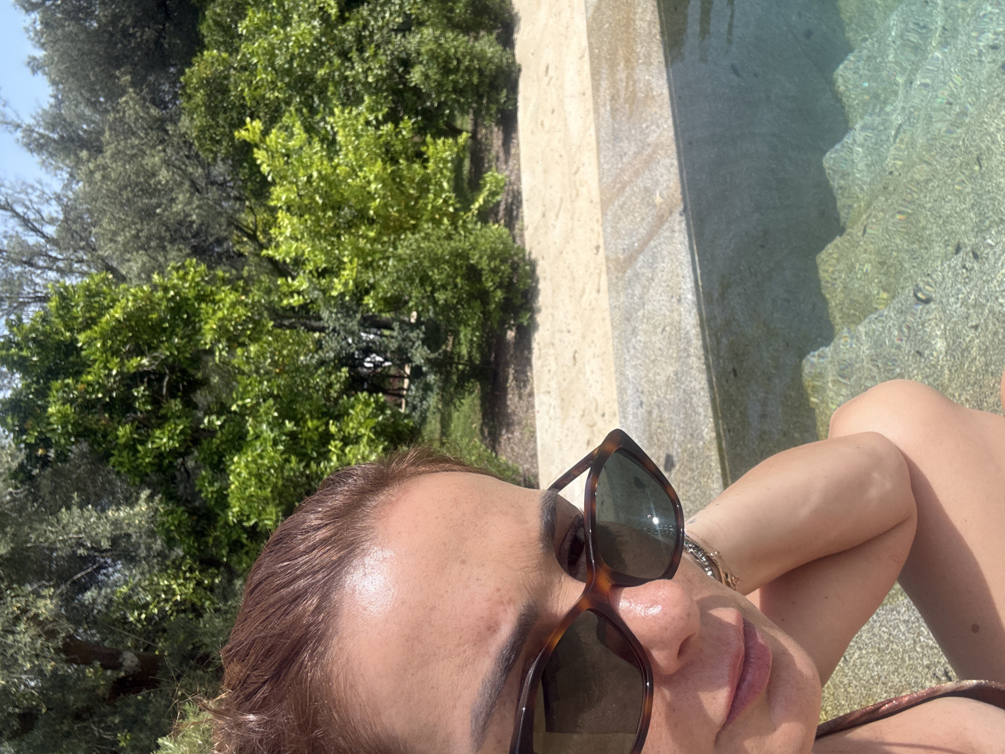 A close-up, tilted selfie photograph shows a person wearing sunglasses and a swimsuit, likely sitting poolside. The person is positioned in the lower portion of the frame. The sunglasses are brown and partially obscure their face. Parts of their shoulder and leg are visible. To the left of the person, there are green trees and shrubbery. In the upper right, there's a light-colored stone edge beside the water, and the water in the pool has a textured appearance. Based on the location provided, the scene is assumed to be somewhere in or around Vila Verde, Portugal, where the subject is presumably relaxing by a swimming pool.