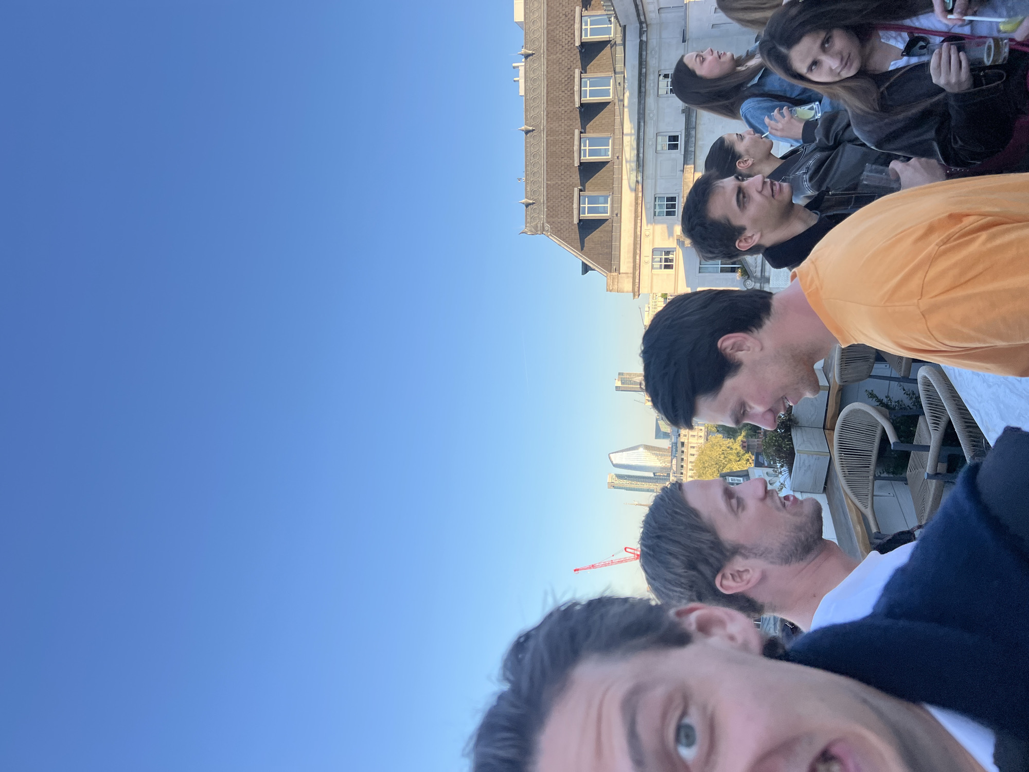 A group of men and women are gathered on a rooftop in London, UK. The background includes a building with multiple windows and a roof, as well as views of city structures and a construction crane. The sky is clear and blue. The people are seemingly engaged in conversation and holding drinks. There are chairs and possibly tables present on the rooftop.