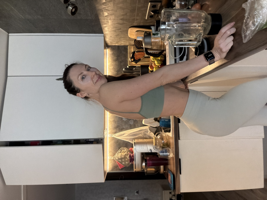 A woman in a green sports bra and leggings stands in a modern kitchen. She leans on a countertop with a large blender. The kitchen features white cabinets and wood-like countertops, with backsplash lighting adding a warm glow. A smart watch is visible on her wrist. She wears eye patches. The setting appears to be a home environment.