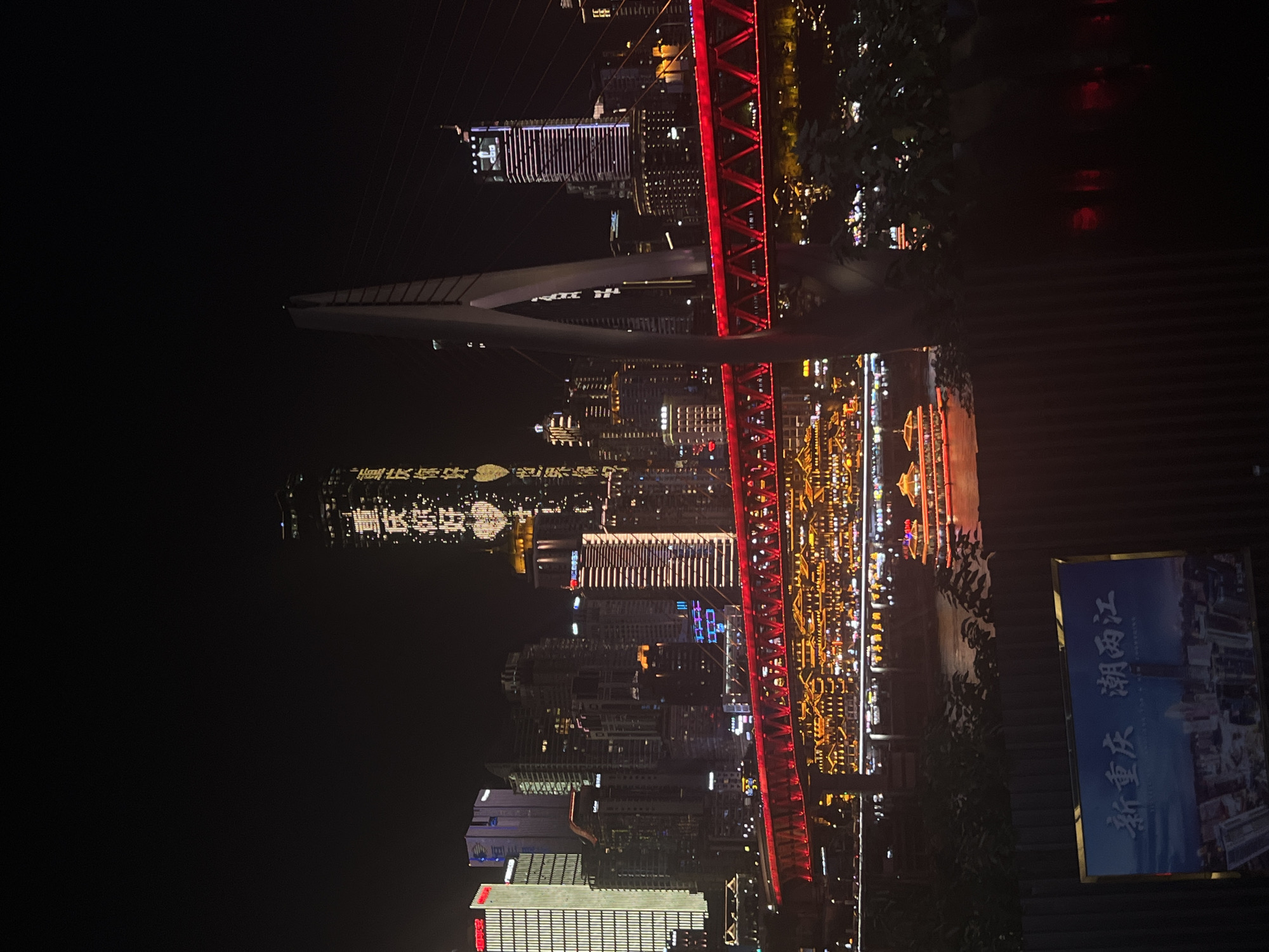 This image depicts a nighttime cityscape, likely in China given the "江北城街道" location context. The scene is dominated by illuminated skyscrapers and a prominent red bridge structure that spans across a body of water, which reflects the city lights. The bridge's trusses are brightly lit in red, creating a striking contrast against the dark sky.
The buildings in the background are a mix of modern architecture, with many featuring elaborate lighting displays. Some buildings are outlined with white and blue lights, while others have rows of warm, yellow lights. The reflection of the city on the water adds depth and a sense of grandeur to the scene.
In the foreground, to the right, a blue sign with white Chinese characters is visible. The text on the sign reads "新重庆 潮两江" (Xīn Chóngqìng Cháo Liǎng Jiāng), which translates to "New Chongqing, Tide of Two Rivers." This text provides a direct location cue and suggests that the image captures a view of Chongqing, China, specifically highlighting its rivers.
The overall mood is one of urban vibrancy and modern development. The image appears to be taken on a clear night, with no visible indications of rain or fog. The time of day is clearly night, indicated by the darkness of the sky and the prevalence of artificial lighting. The scene evokes a sense of awe and wonder at the scale and beauty of the illuminated city. There are no visible people in the image.